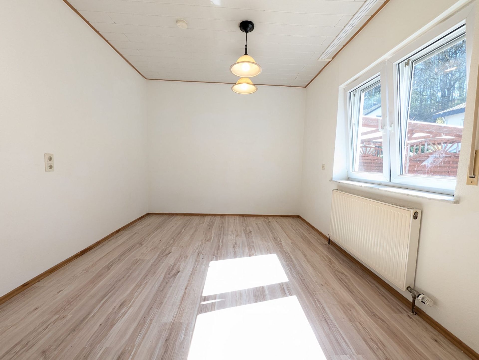 Heller, leerer Raum mit Holzfußboden, zwei Fenstern und Deckenlampe.