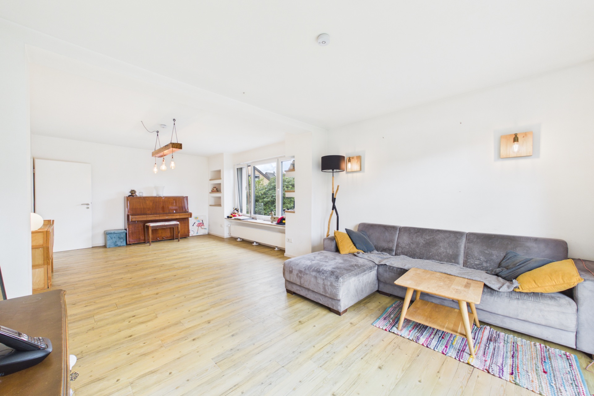 Helles Wohnzimmer mit grauem Sofa, Holztisch, Klavier und großen Fenstern.