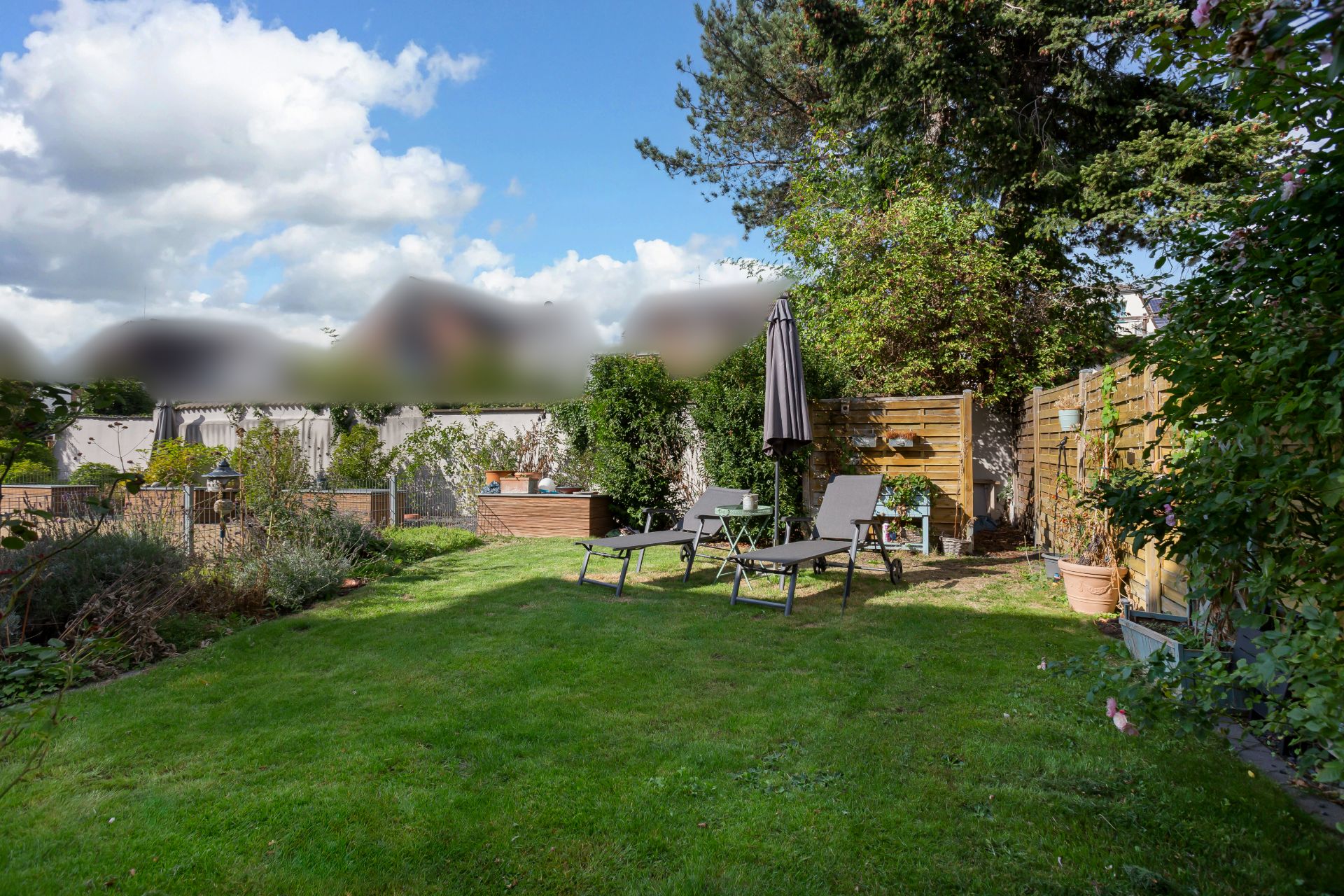 Garten mit Liegestühlen, Sonnenschirm und viel Grün, umgeben von Holzzäunen und Pflanzen.