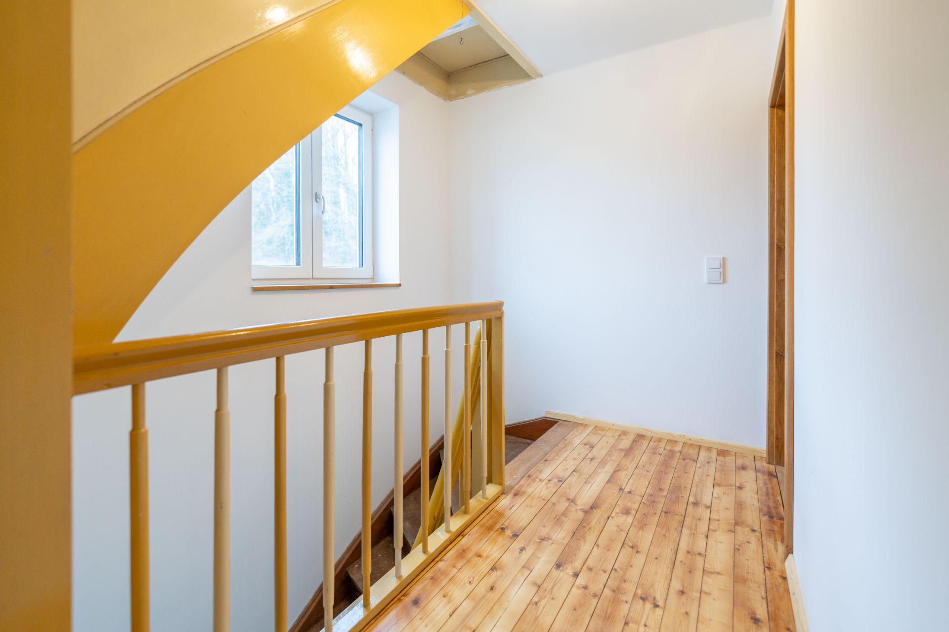 Heller Flur mit Holzgeländer, Fenster und Treppe, Holzboden und weißen Wänden.