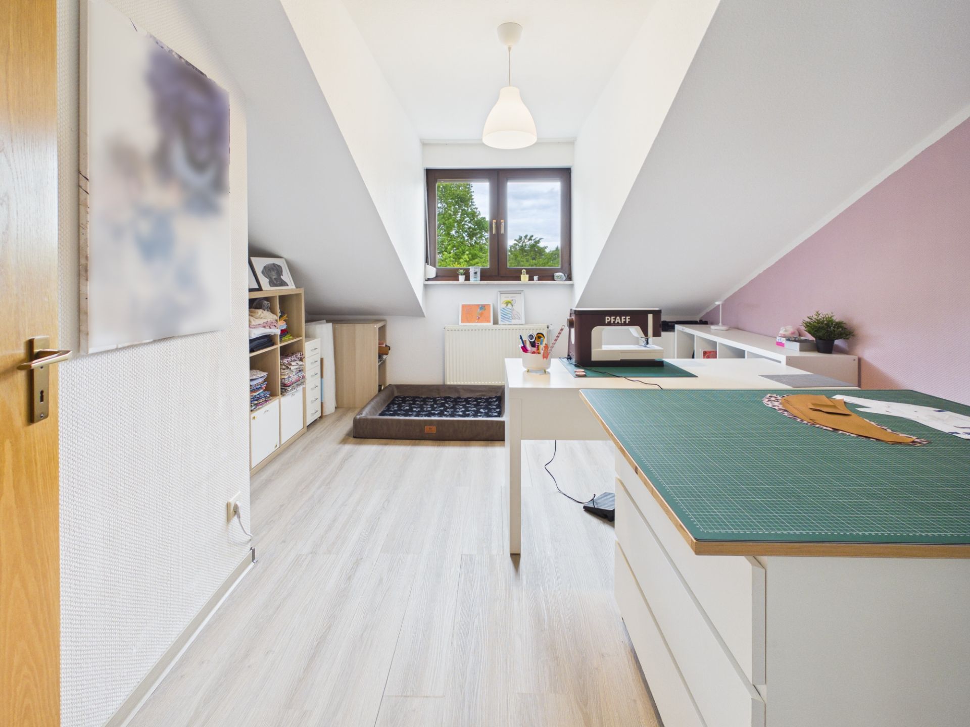 Helles Arbeitszimmer mit Schreibtisch, Regalen und Nähmaschine unter Dachschrägen.