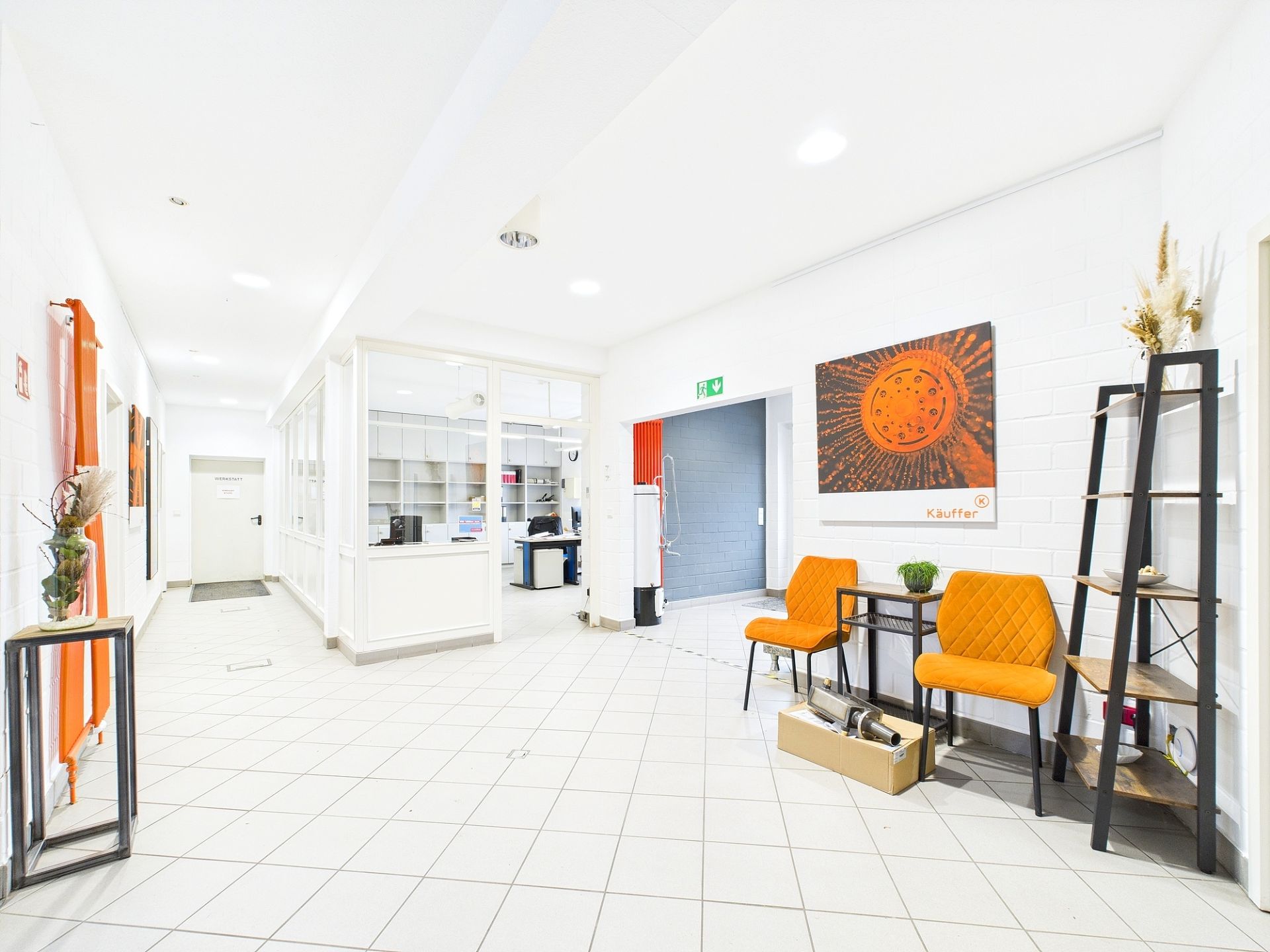 Heller Flur mit zwei orangefarbenen Stühlen, Wandbild und Regal, daneben ein Büro hinter Glaswand.