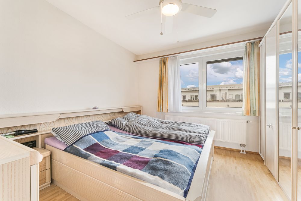 Helles Schlafzimmer mit Doppelbett, Decke im Karomuster und großem Fenster mit Blick nach draußen.