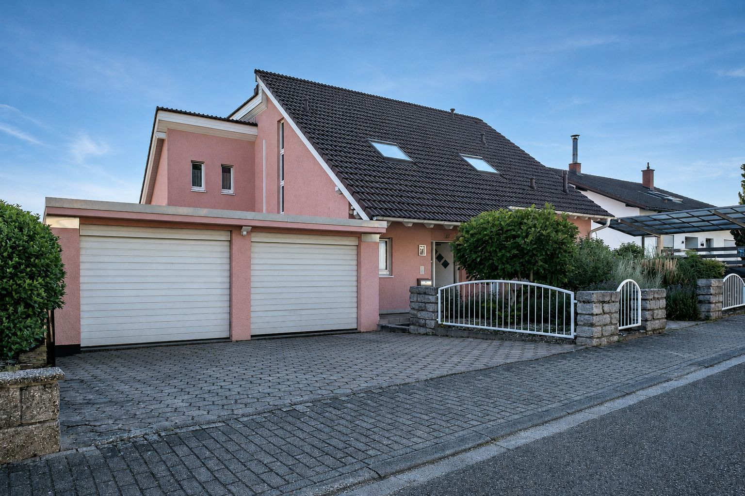 Modernes Einfamilienhaus mit rosa Fassade, zwei Garagen und Vorgarten an einer Straße.