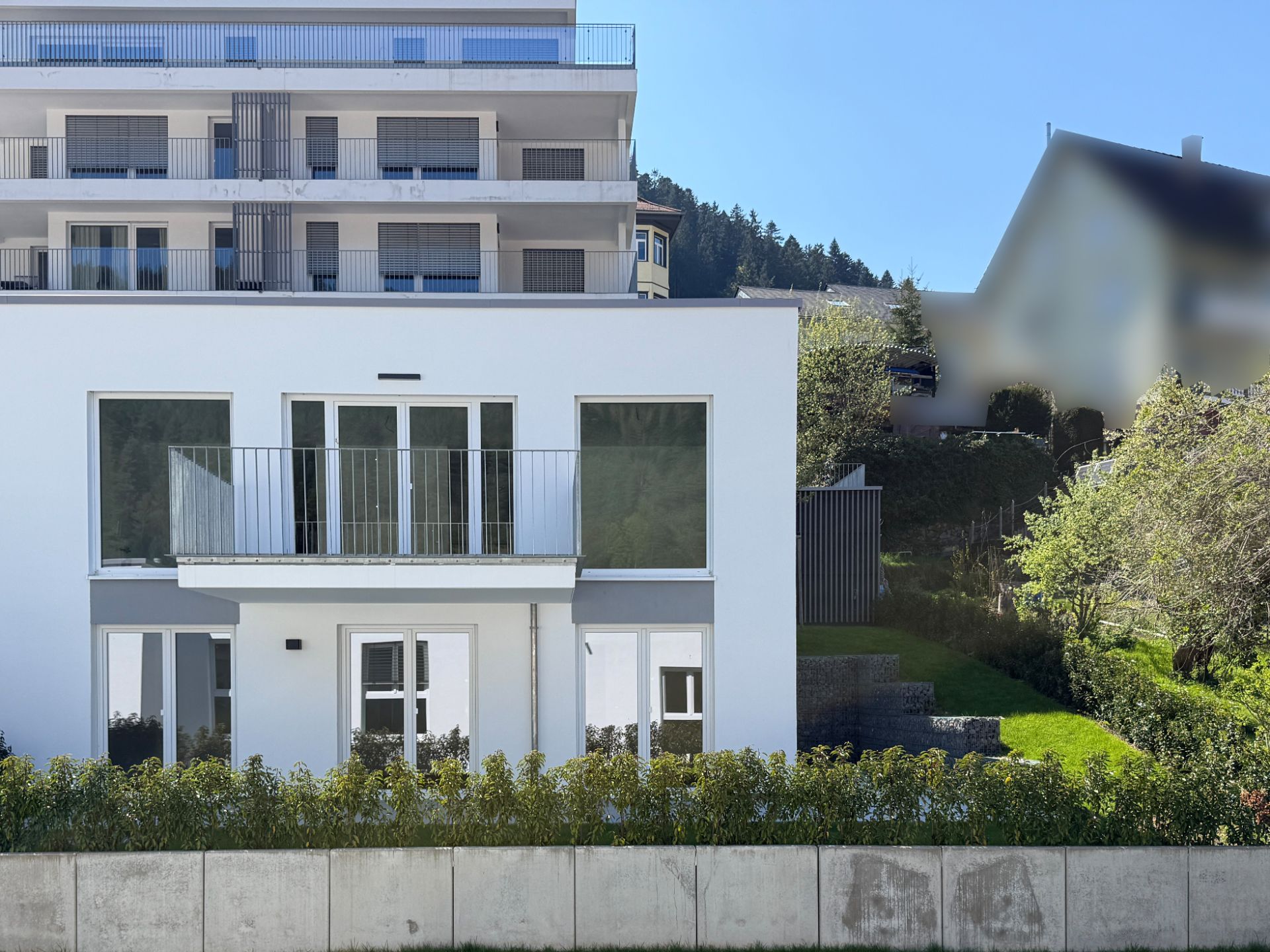 Modernes weißes Wohnhaus mit Balkon und großen Fenstern, davor Hecke und Garten.