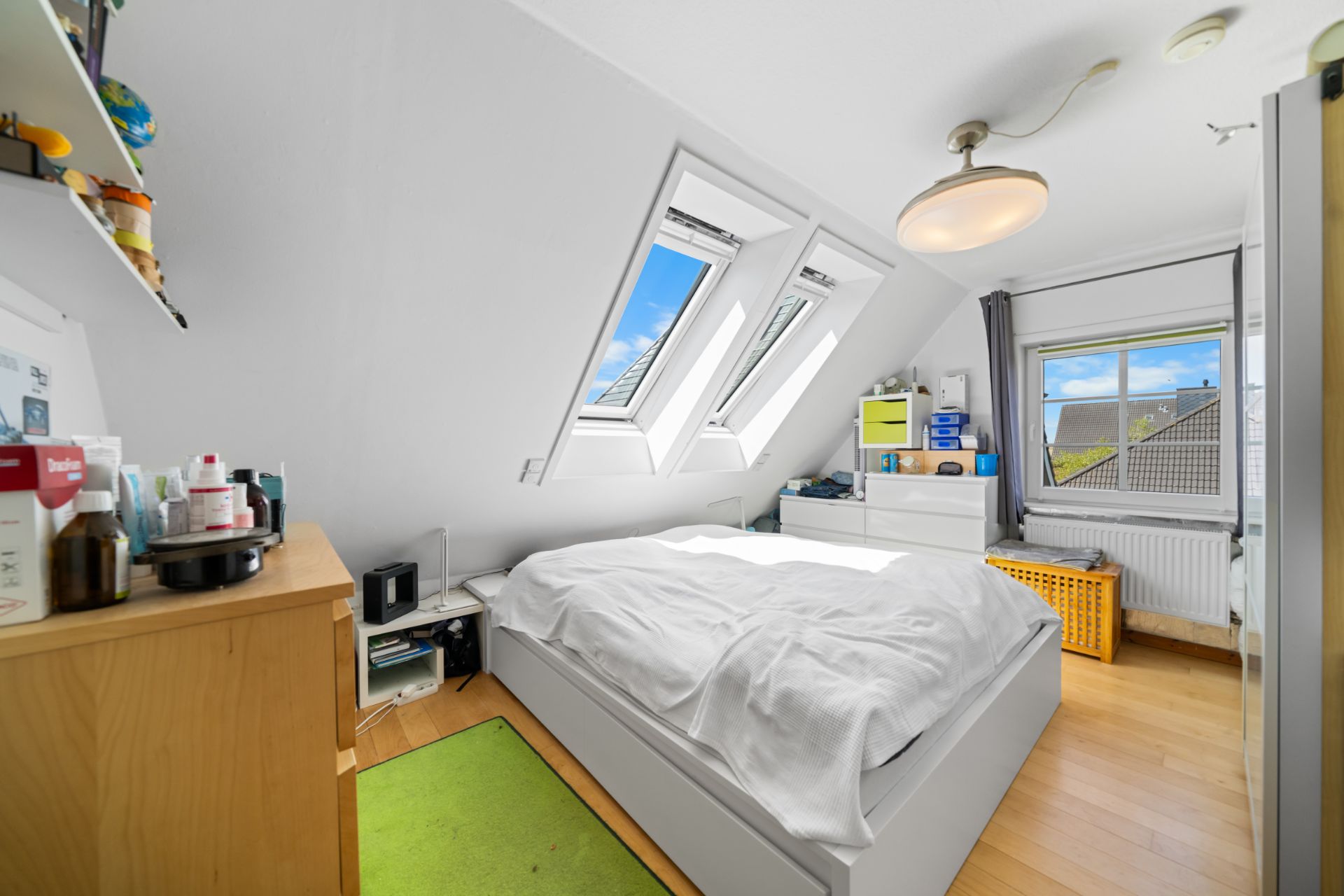 Helles Schlafzimmer mit Dachfenstern, Bett, Kommode und grünem Teppich auf Holzfußboden.