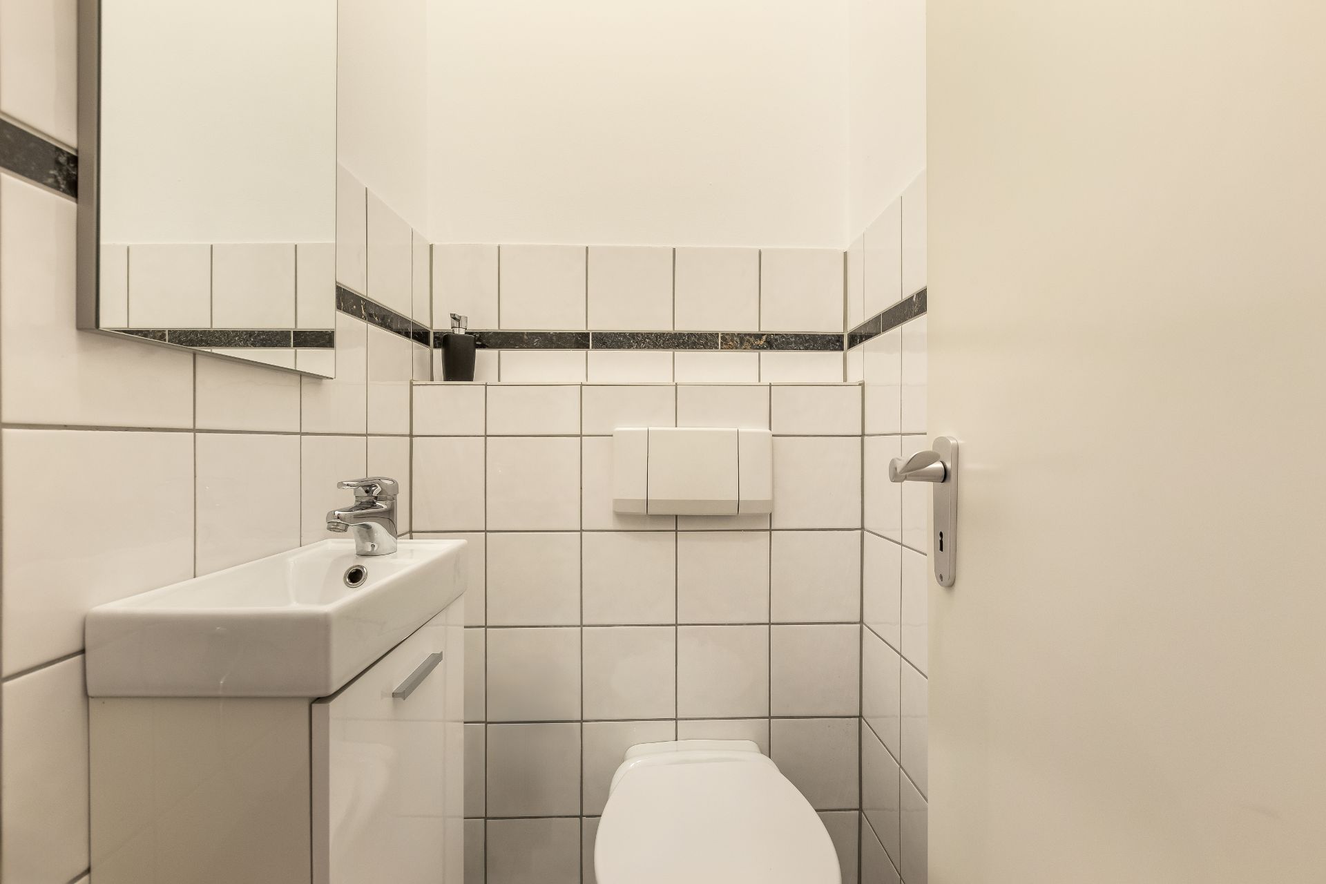 Kleines Badezimmer mit Toilette, Waschbecken und Wandspiegel, weiße Fliesen an den Wänden.
