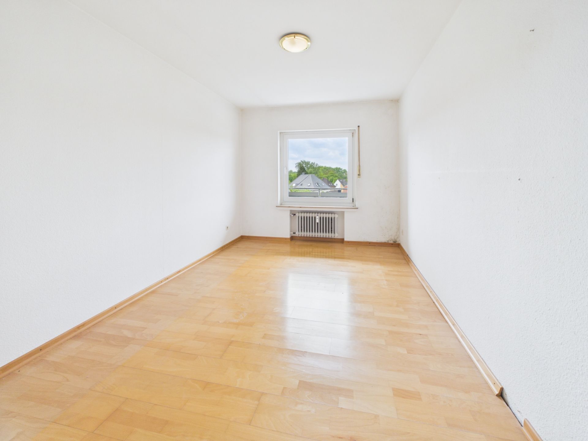 Leerer, heller Raum mit Holzfußboden, weiß gestrichenen Wänden und Fenster mit Blick nach draußen.