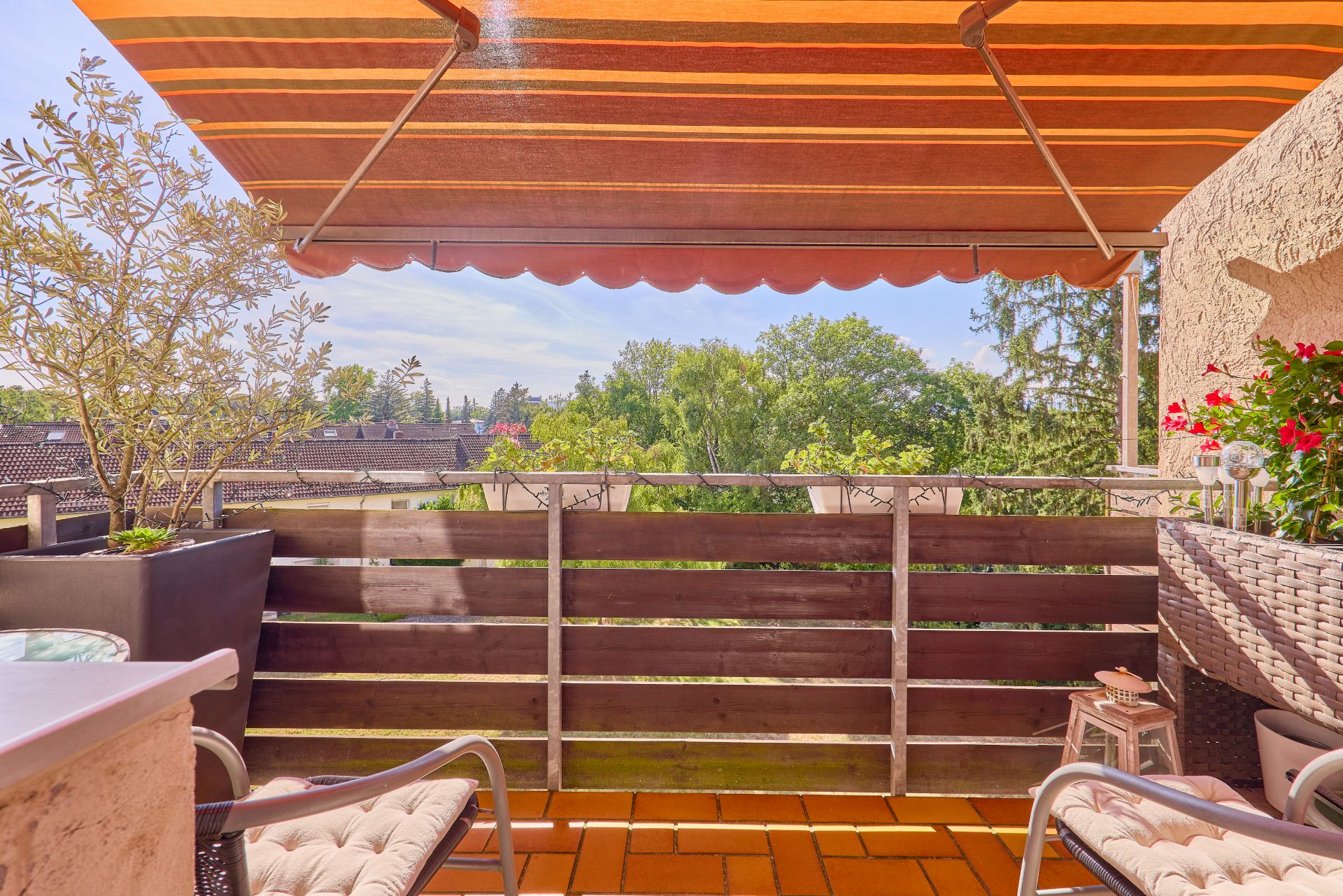 Balkon mit Markise, Pflanzen, zwei Stühlen und Blick auf Bäume und Dächer.