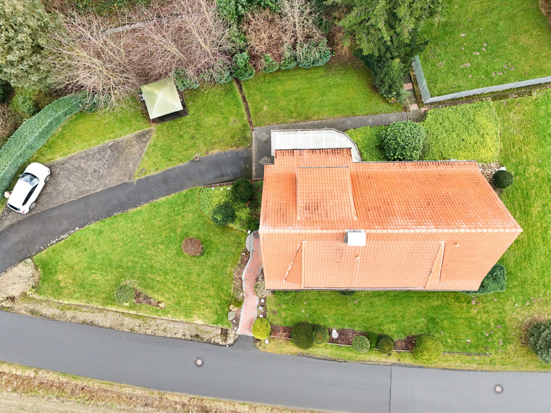 Luftaufnahme eines Hauses mit rotem Dach, Auffahrt, Auto und gepflegtem Garten.