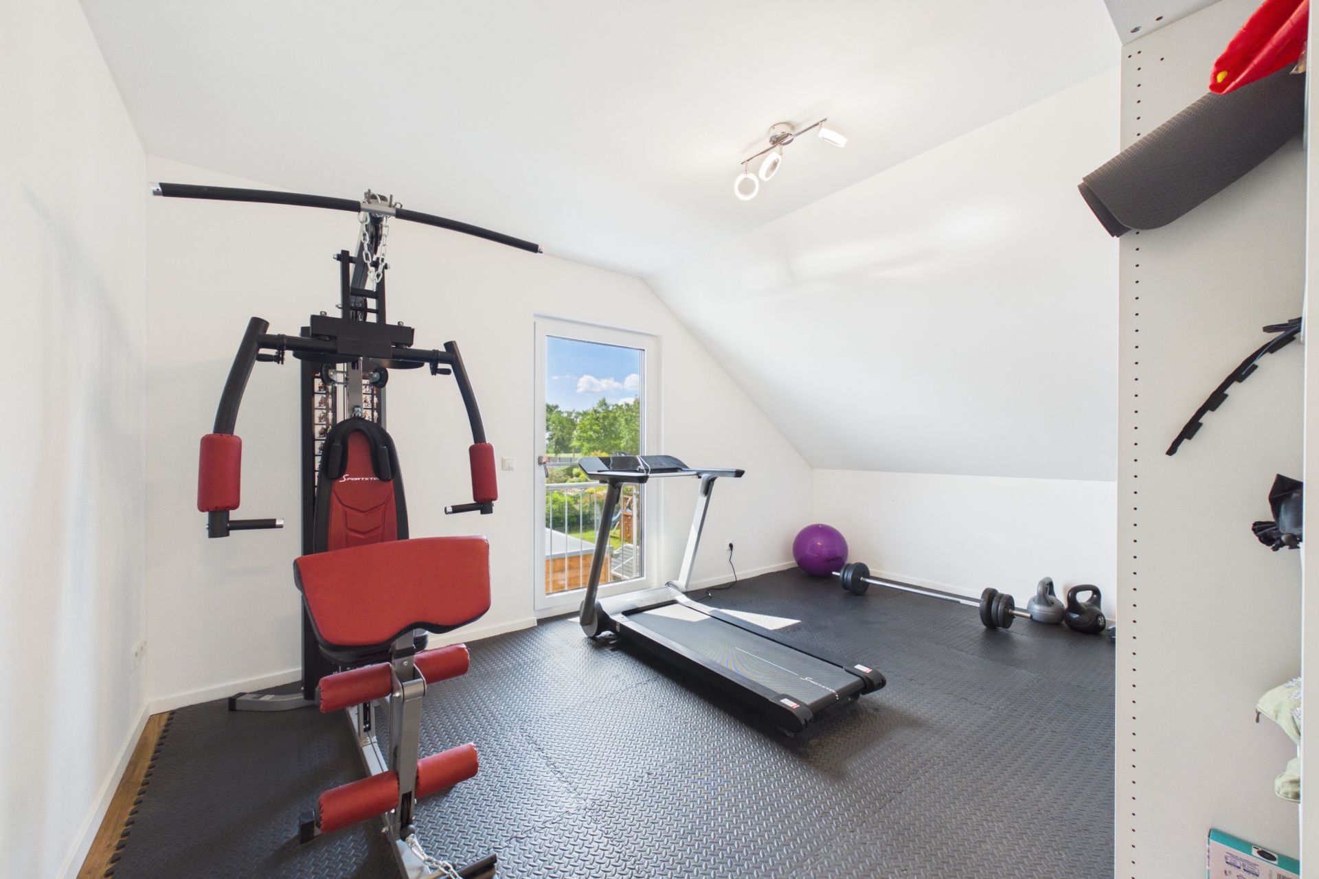 Heller Fitnessraum mit Trainingsgerät, Laufband, Hanteln und Gymnastikball.