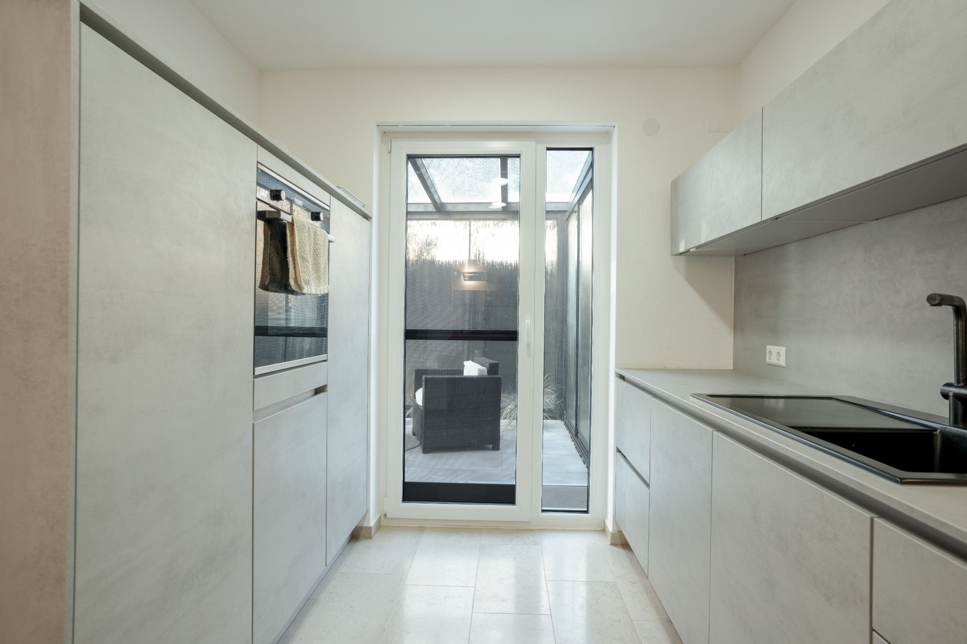 Moderne Küche mit hellen Schränken, Backofen und Zugang zu einer Terrasse mit Glasüberdachung.