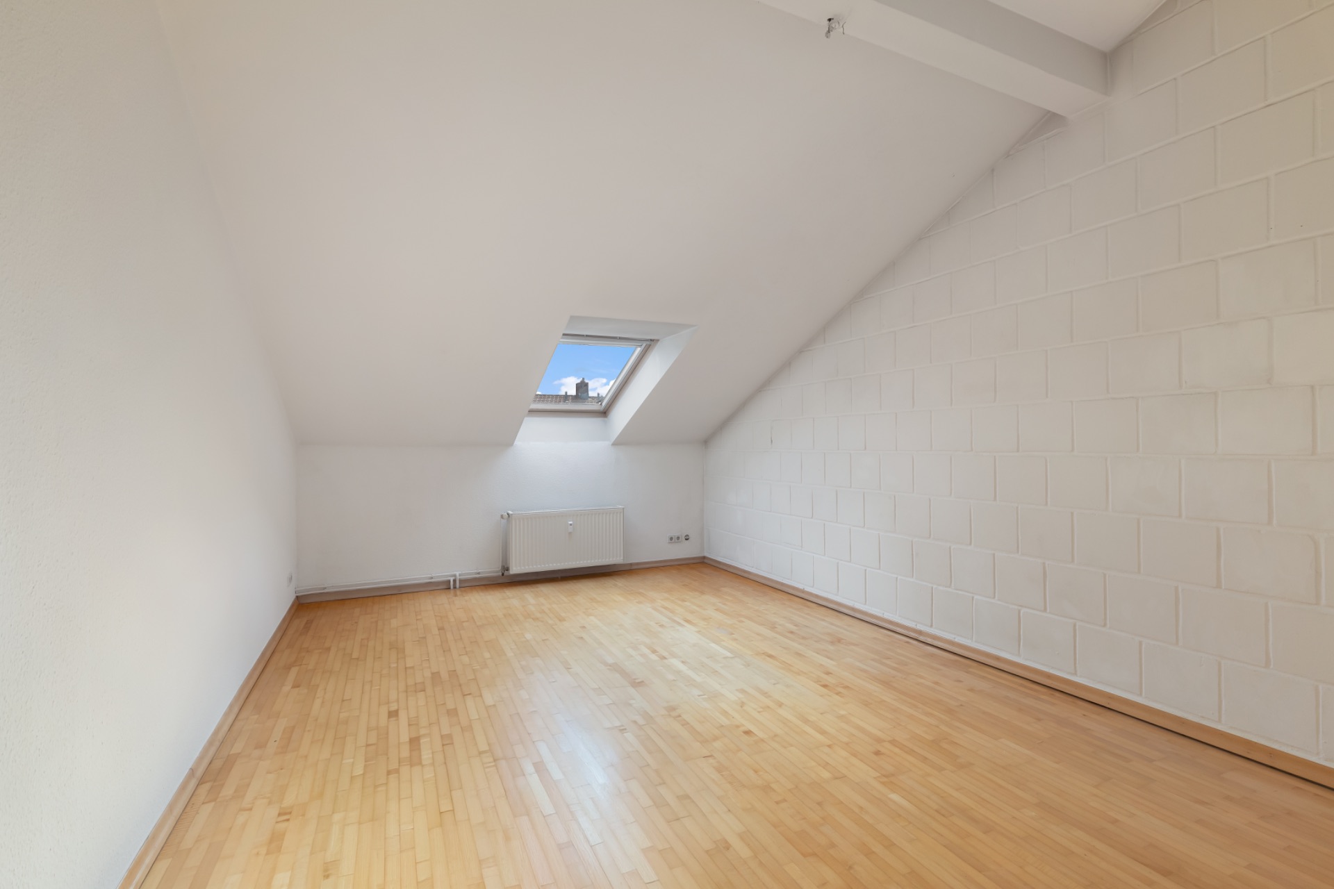 Leerer Raum mit schräger Decke, Dachfenster und hellem Holzboden.