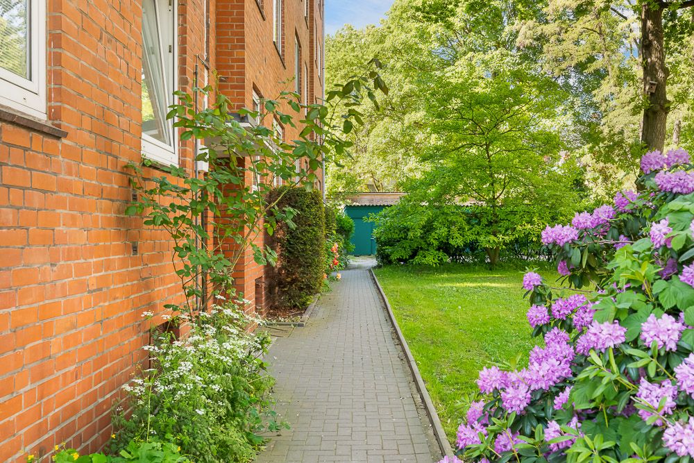 Gepflasterter Weg entlang einer roten Hauswand mit Blumengarten und grüner Wiese.