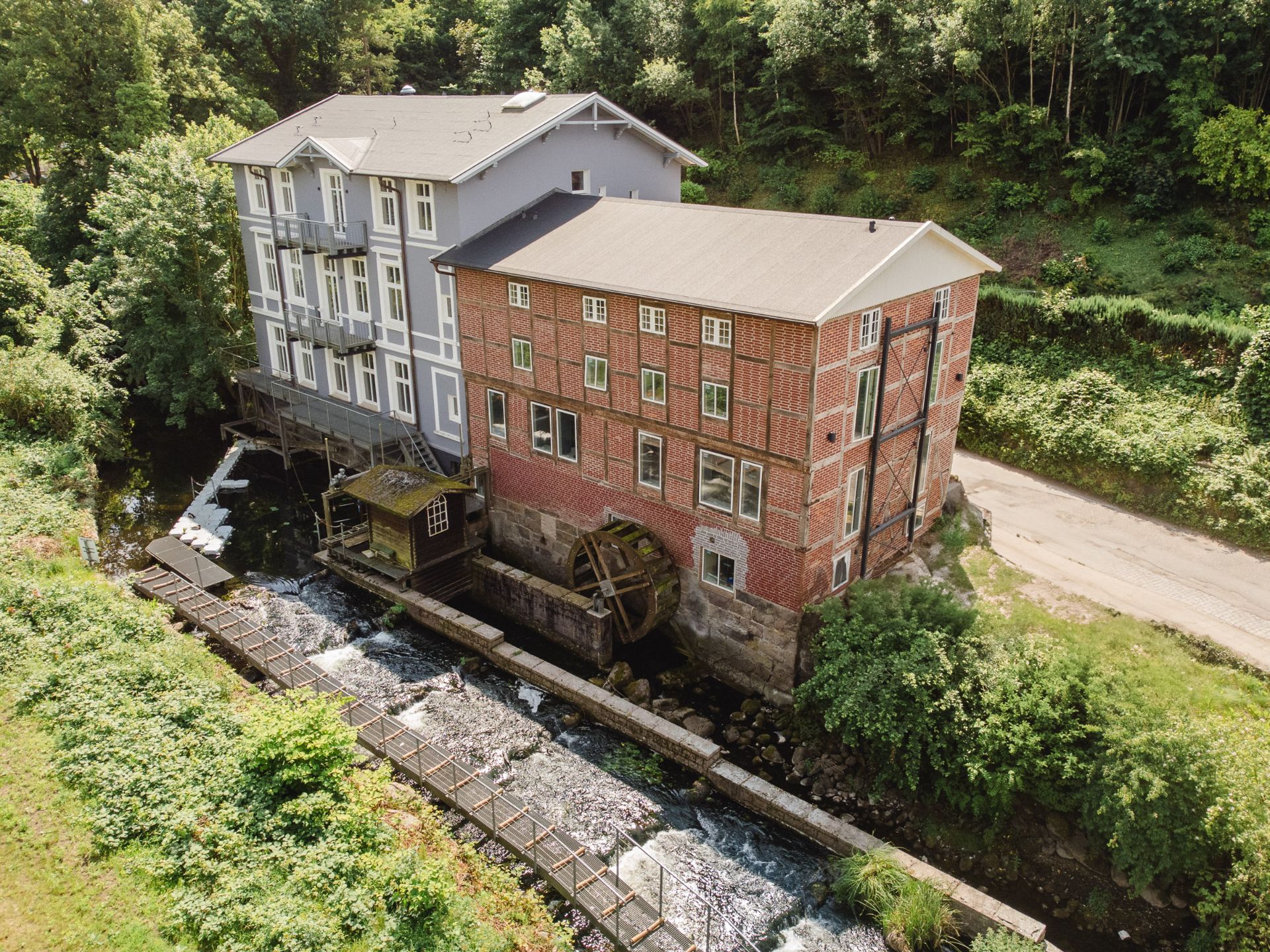 Zwei Wohngebäude an einem Bach, eines aus Backstein mit Wasserrad, umgeben von Bäumen und Grün.