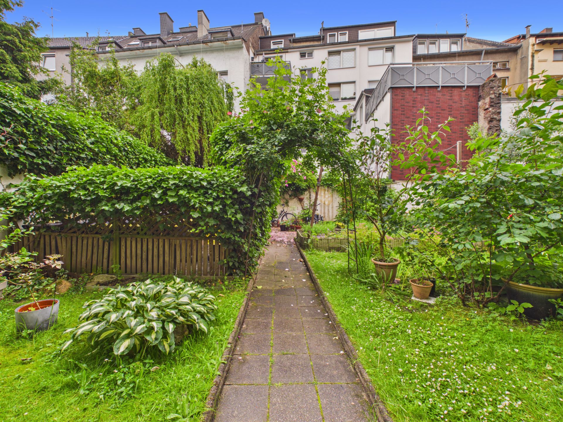 Garten mit gepflastertem Weg, Pflanzen und Blumen, umgeben von Wohnhäusern.