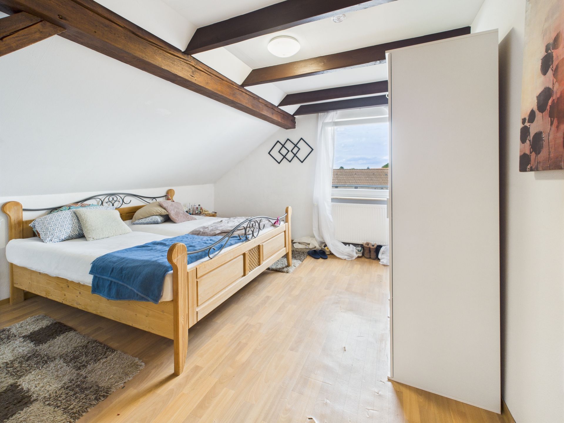 Schlafzimmer mit Doppelbett, Holzboden, weißem Schrank und Fenster unter schrägem Dach.