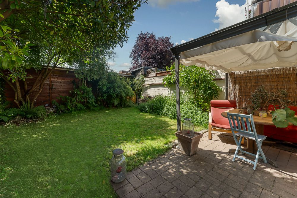 Garten mit Rasenfläche, Bäumen und überdachter Terrasse mit Sitzgruppe und Tisch.