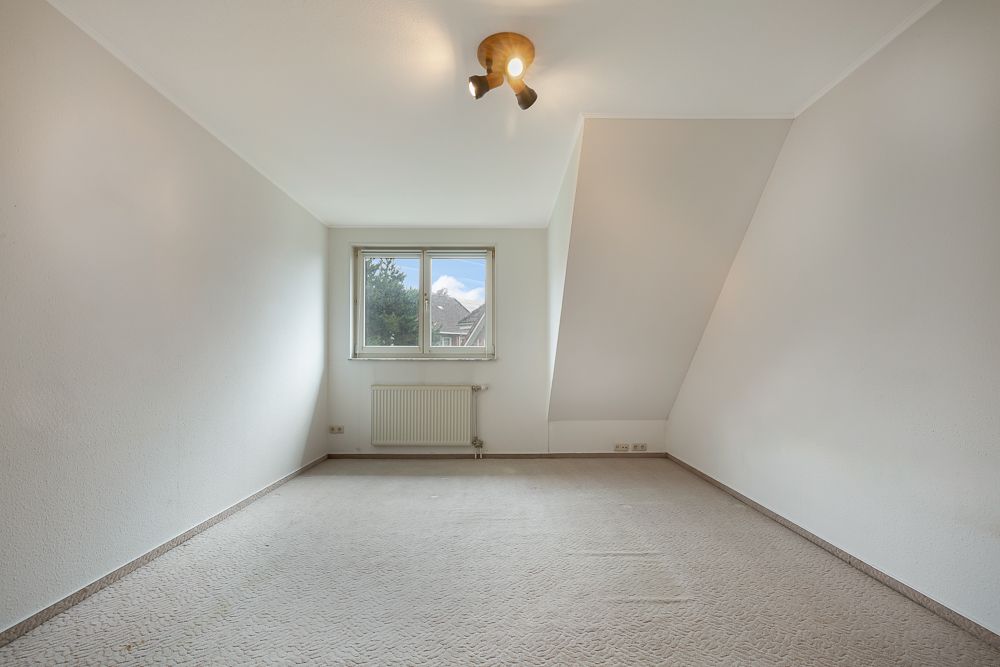 Leeres Zimmer mit Teppichboden, Fenster, Heizkörper und abgeschrägter Wand.
