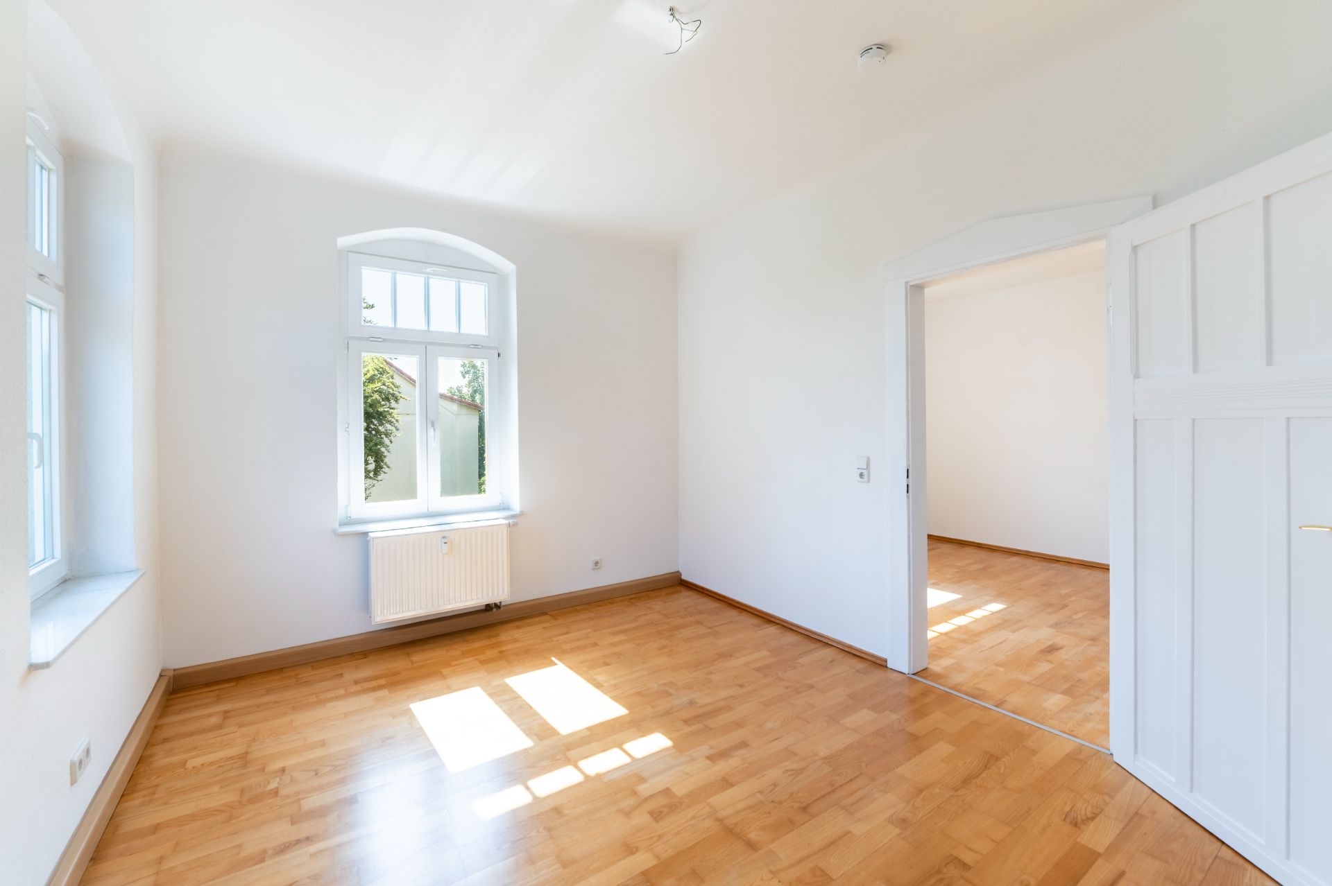 Heller Raum mit Parkettboden, weißen Wänden und drei Fenstern, verbunden mit einem weiteren Zimmer.