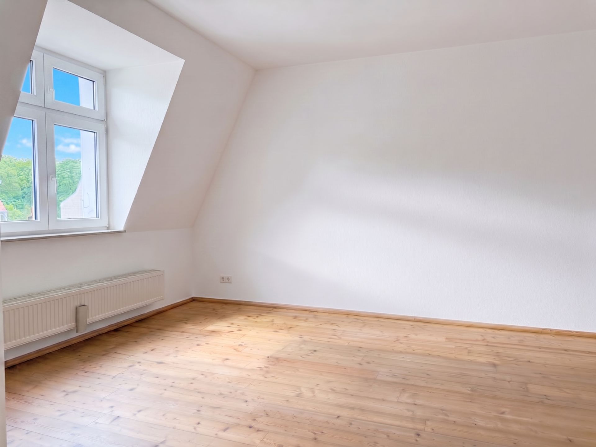 Helles, leeres Zimmer mit Holzfußboden, weißer Wand und großem Fenster.