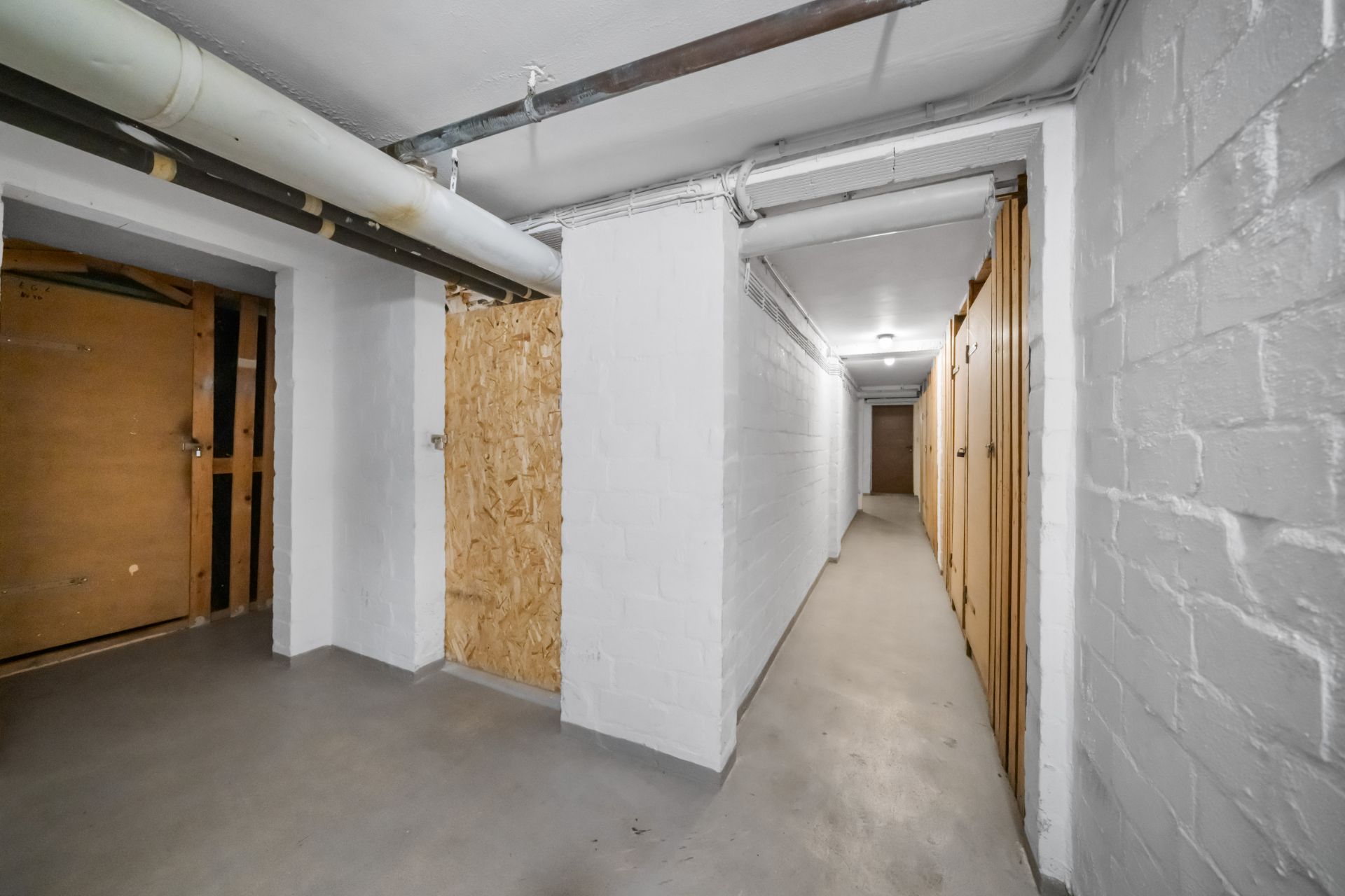 Flur im Keller mit weißen Wänden, Türen und sichtbaren Rohren an der Decke.
