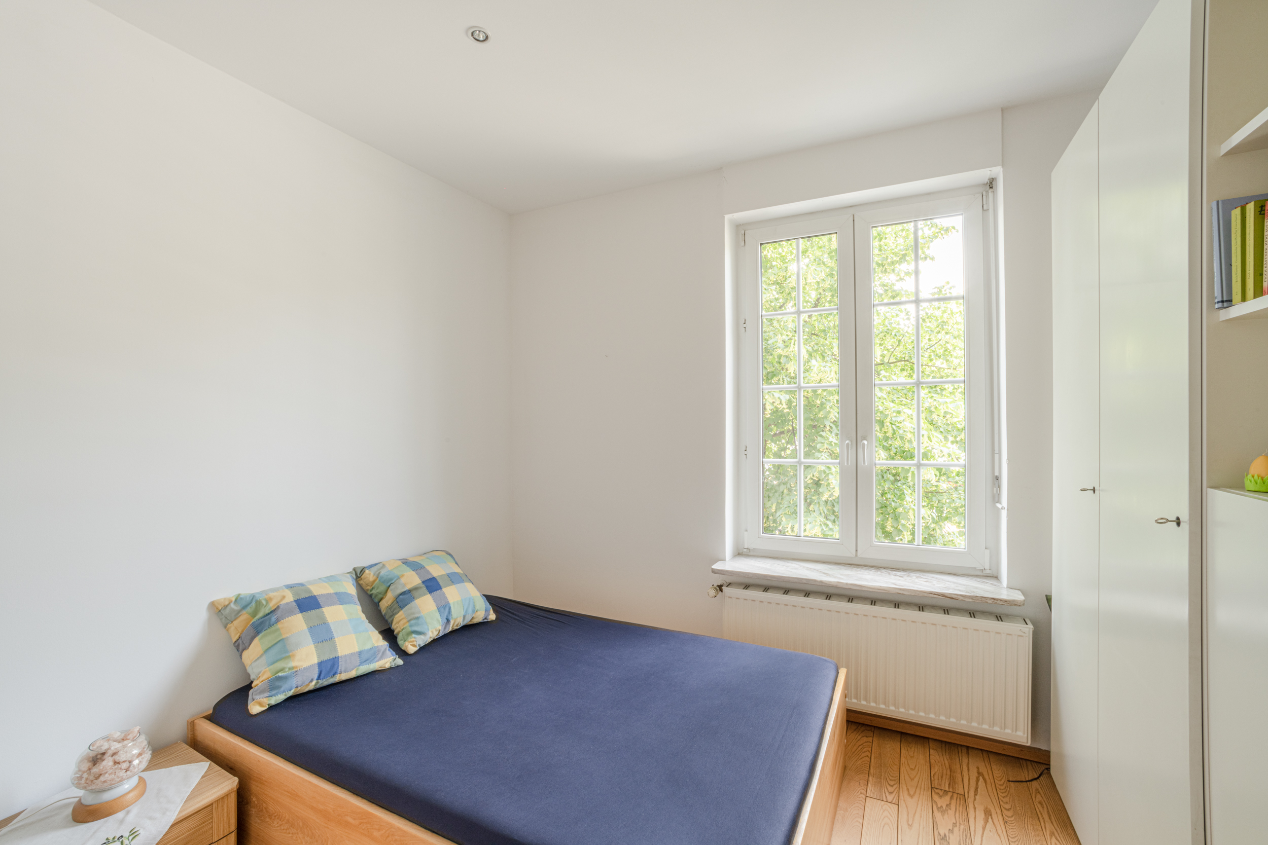 Helles Schlafzimmer mit Bett, Nachttisch, großem Fenster und weißem Schrank.