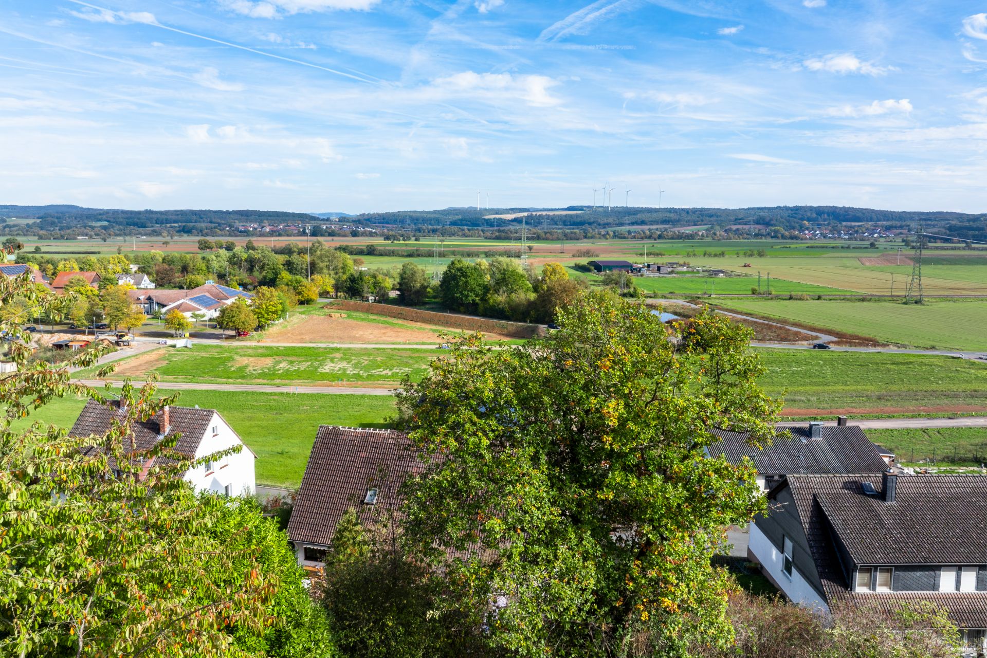 Blick auf Felder, Bäume und Wohnhäuser in ländlicher Umgebung bei klarem Himmel.