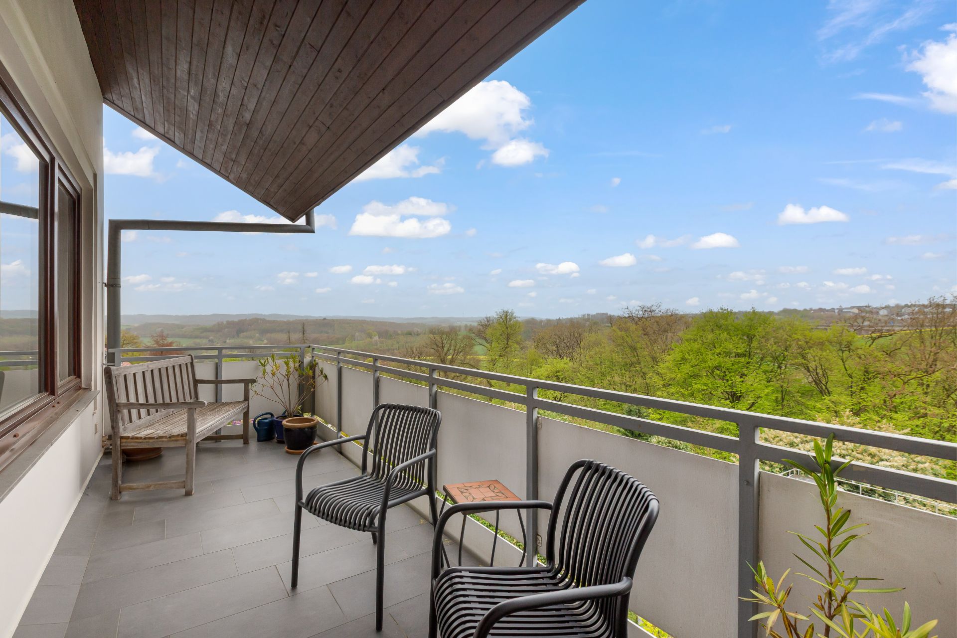 Balkon mit zwei Stühlen, einer Bank, Pflanzen und weitem Ausblick auf grüne Landschaft.