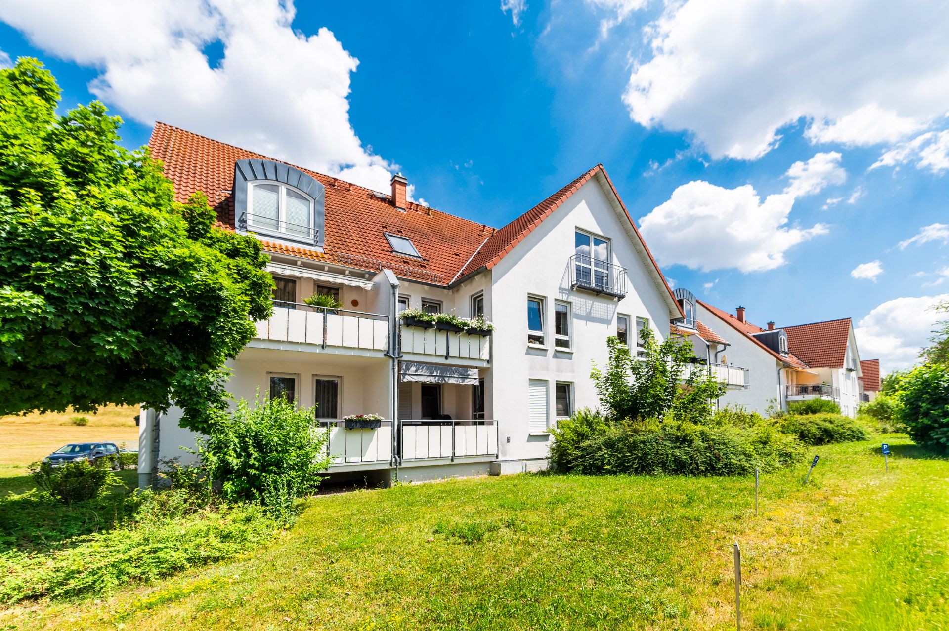 Mehrfamilienhaus mit Balkonen, rotem Dach und grüner Wiese davor bei sonnigem Wetter.