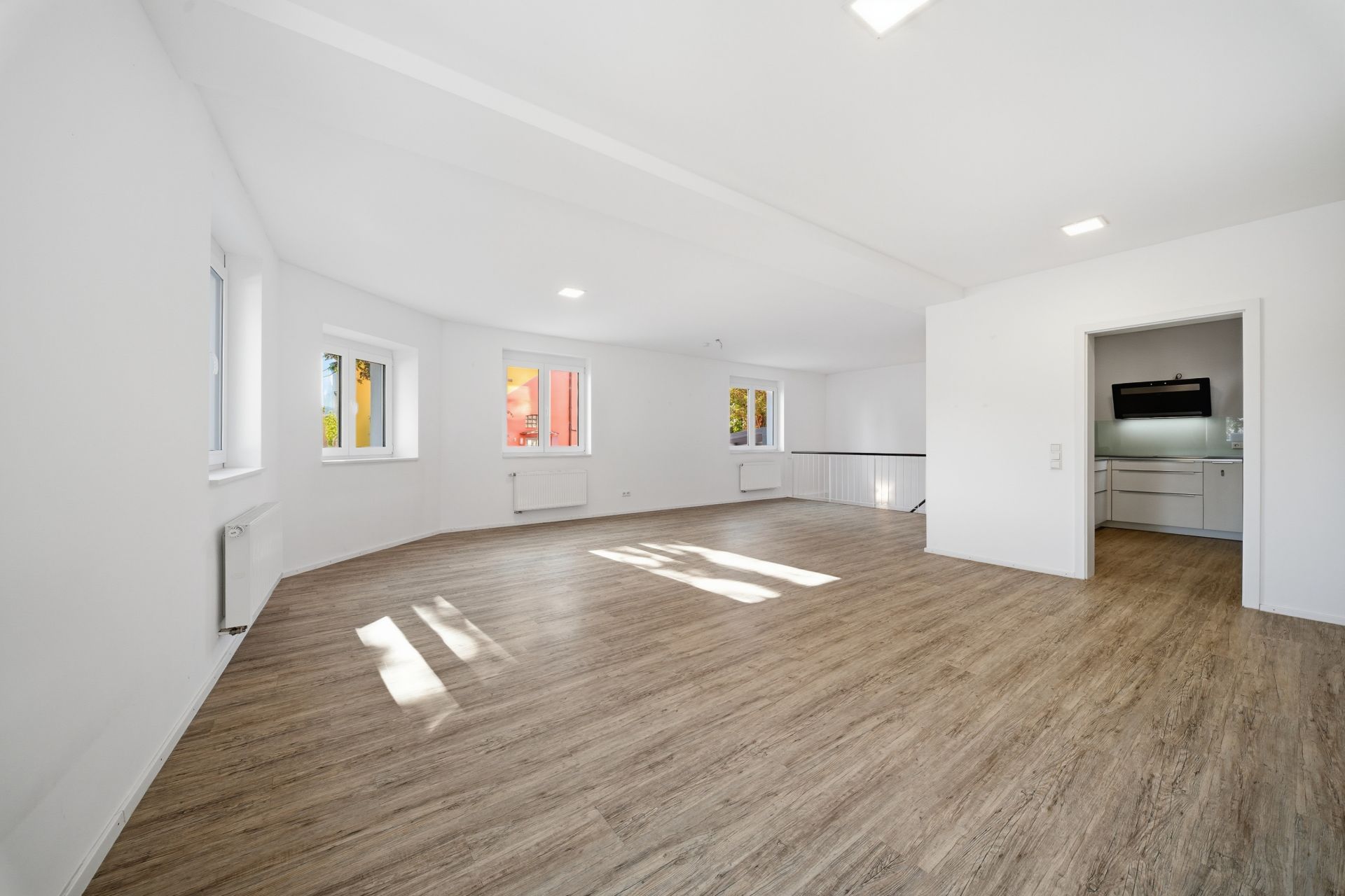 Heller Wohnraum mit Holzfußboden, mehreren Fenstern und angrenzender Küche.