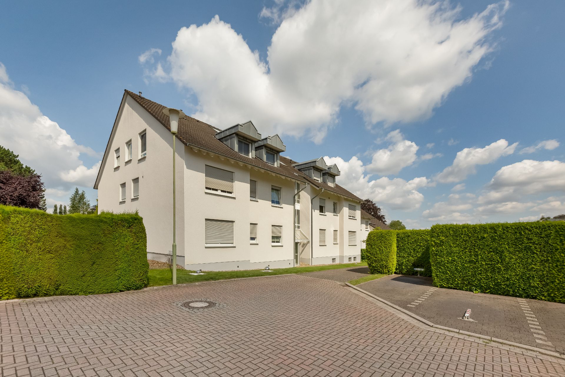 Weißes Wohngebäude mit mehreren Fenstern, umgeben von grünen Hecken und gepflastertem Parkplatz.