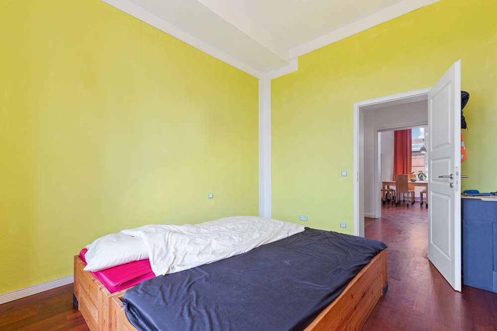 Schlafzimmer mit Holzbett, gelben Wänden und offener Tür zum nächsten Raum.