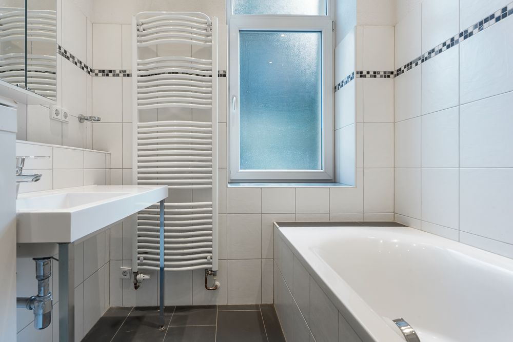 Helles Badezimmer mit Waschbecken, Badewanne, Handtuchheizkörper und Fenster.