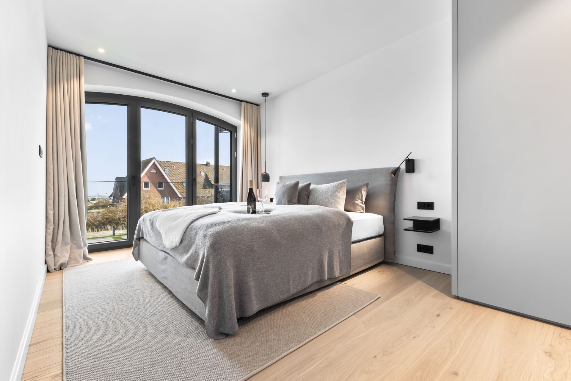Modernes Schlafzimmer mit großem Bett, hellen Vorhängen und bodentiefem Fenster mit Blick nach draußen.