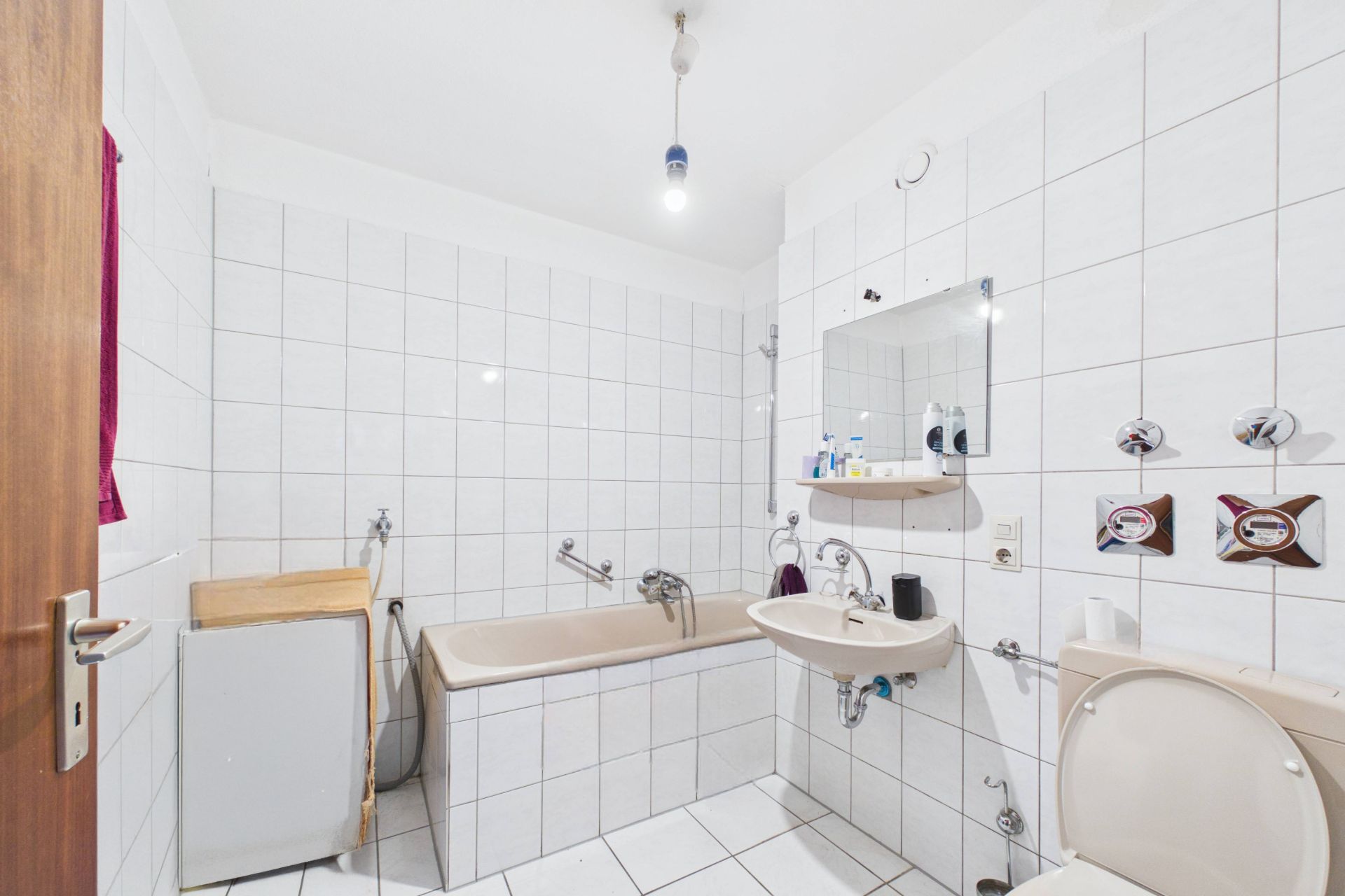 Badezimmer mit Badewanne, Waschbecken, Toilette und Spiegel. Weiße Fliesen an Wänden und Boden.