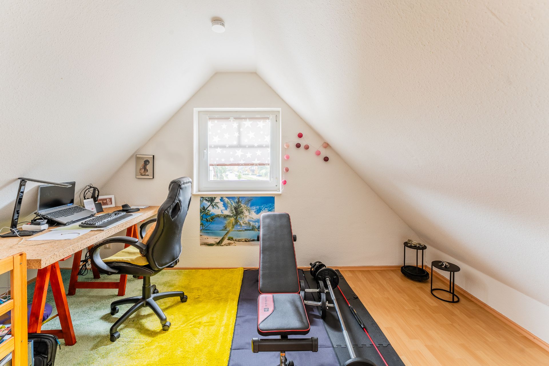 Dachzimmer mit Schreibtisch, Bürostuhl und Hantelbank unter einem Fenster.