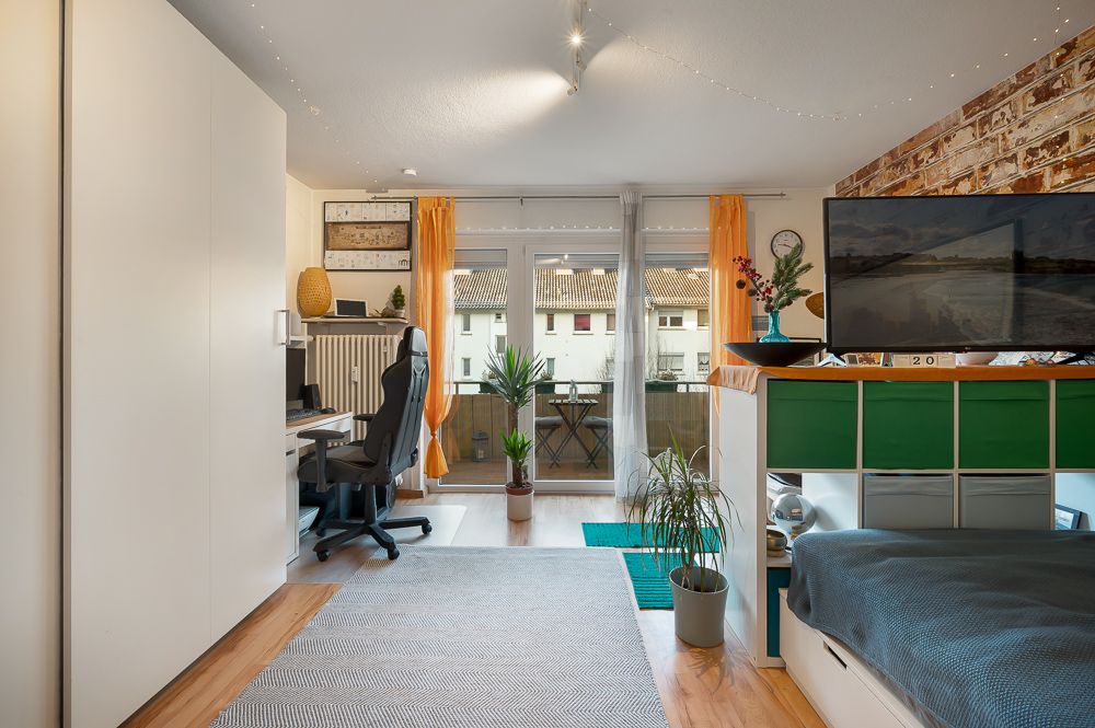 Helles Zimmer mit Bett, Schreibtisch, Fernseher, Pflanzen und Balkonzugang.