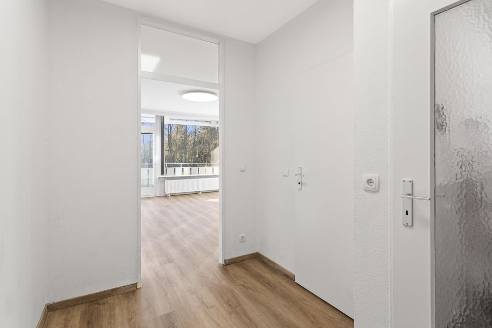 Heller Flur mit Holzboden, weißen Wänden und Blick ins Wohnzimmer mit großen Fenstern.