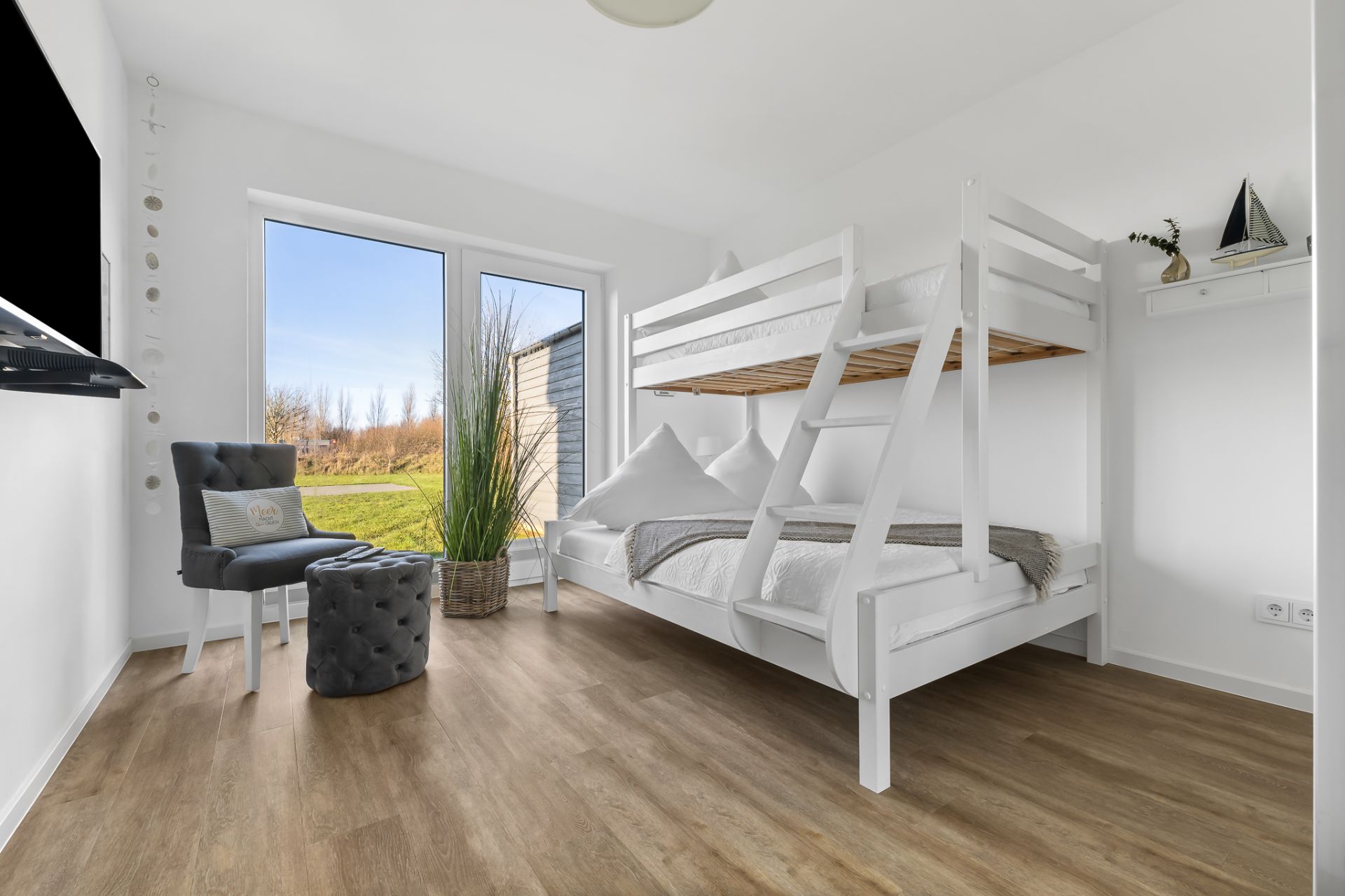 Modernes Schlafzimmer mit Etagenbett, Sessel, Hocker und großem Fenster mit Blick ins Grüne.
