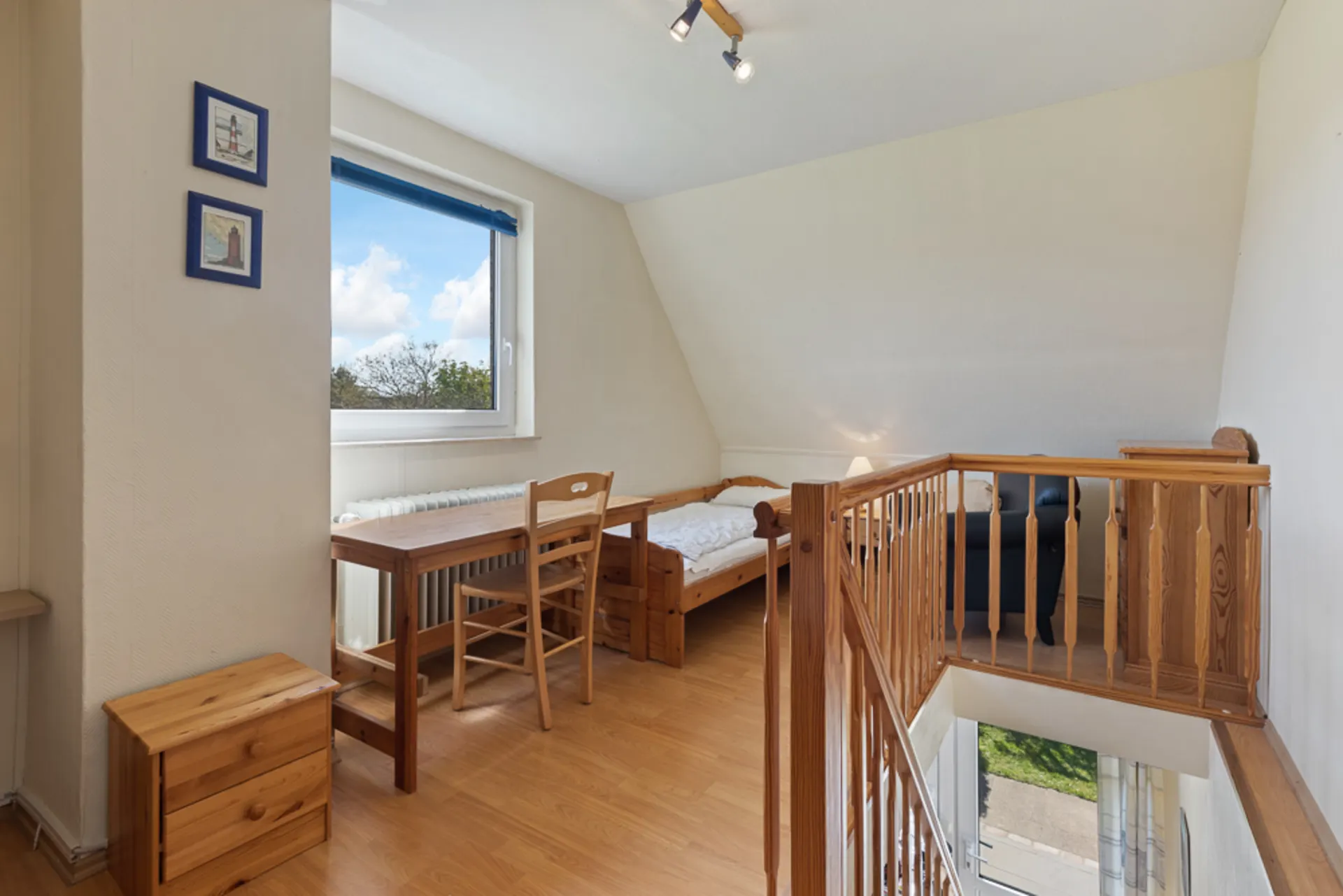Helles Zimmer mit Holztreppe, Einzelbett, Schreibtisch, Stuhl und Fenster mit blauem Rollo.