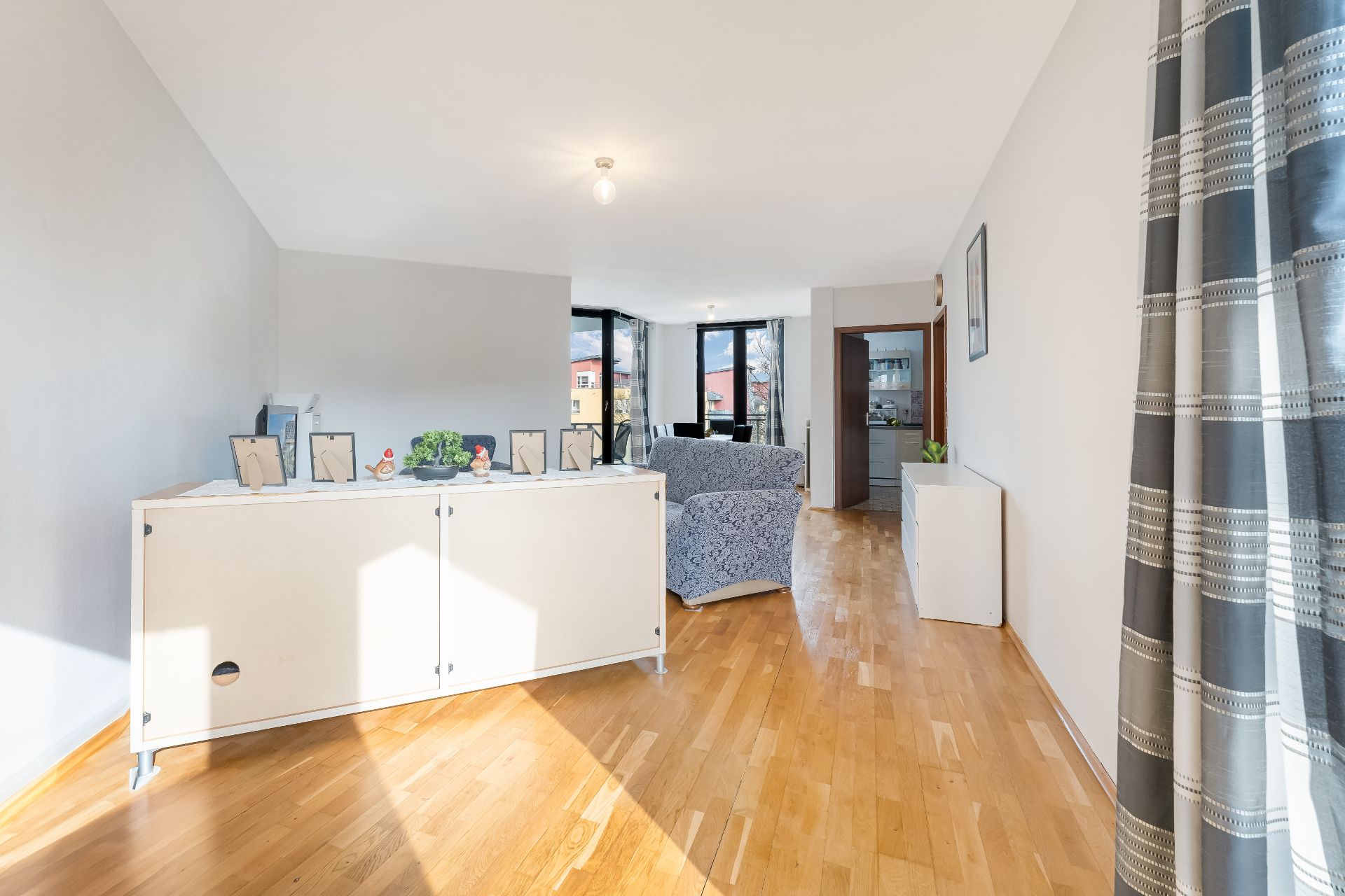 Helles Wohnzimmer mit Parkettboden, Sideboard, zwei Sesseln und großen Fenstern.
