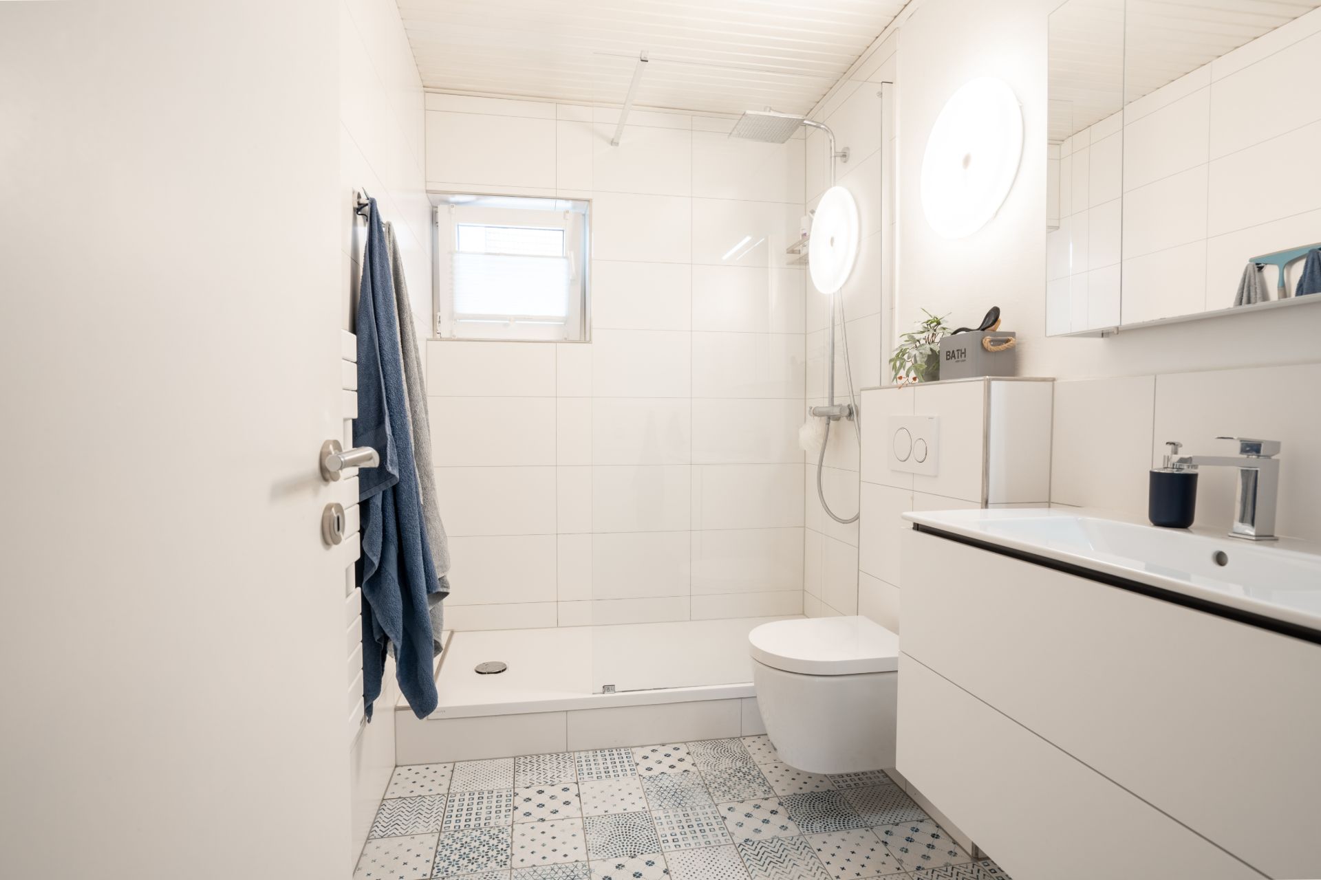 Helles Badezimmer mit Dusche, WC, Waschbecken, Handtüchern und gemusterten Fliesen.
