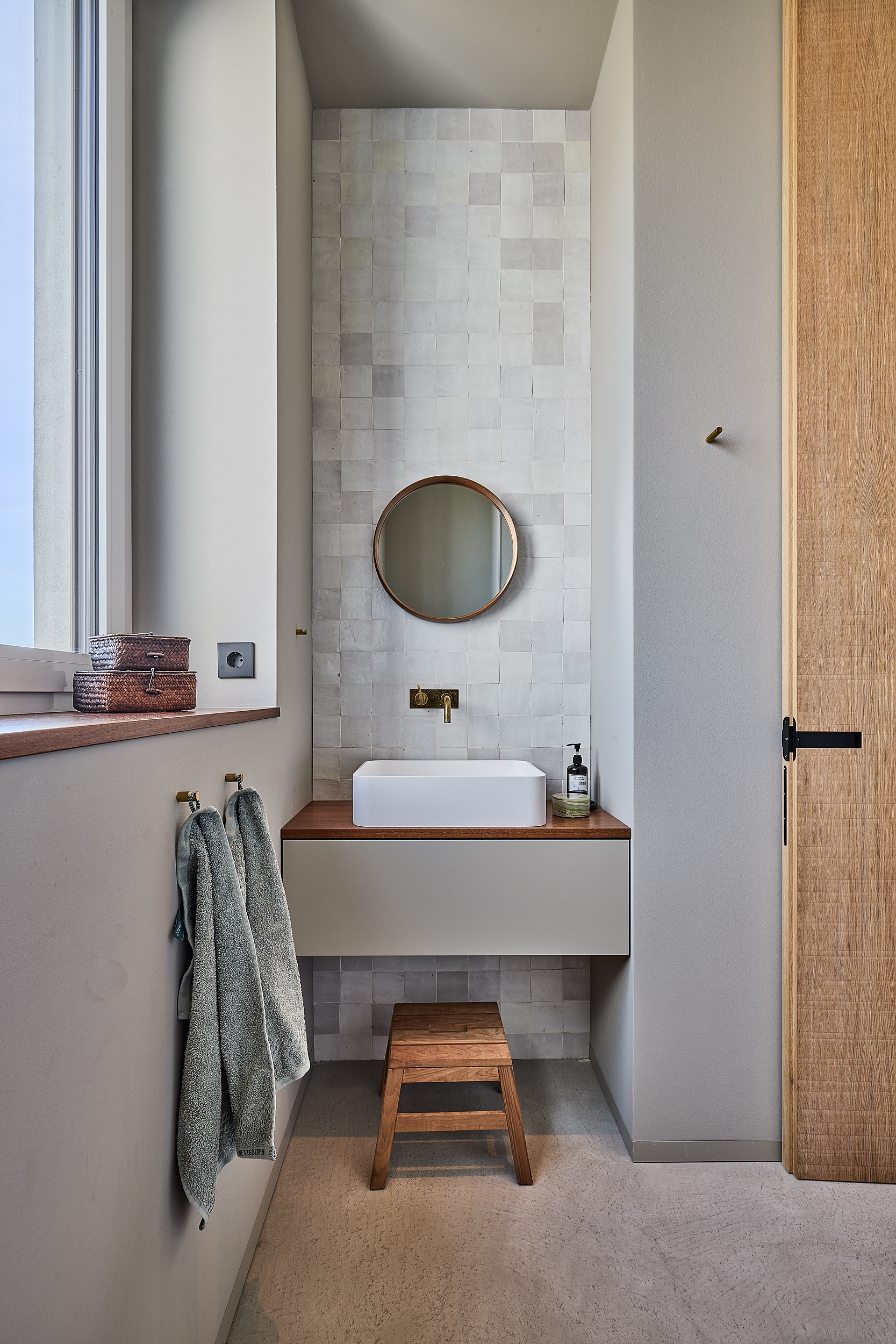 Modernes Badezimmer mit Waschbecken, rundem Spiegel und Hocker vor gefliester Wand.