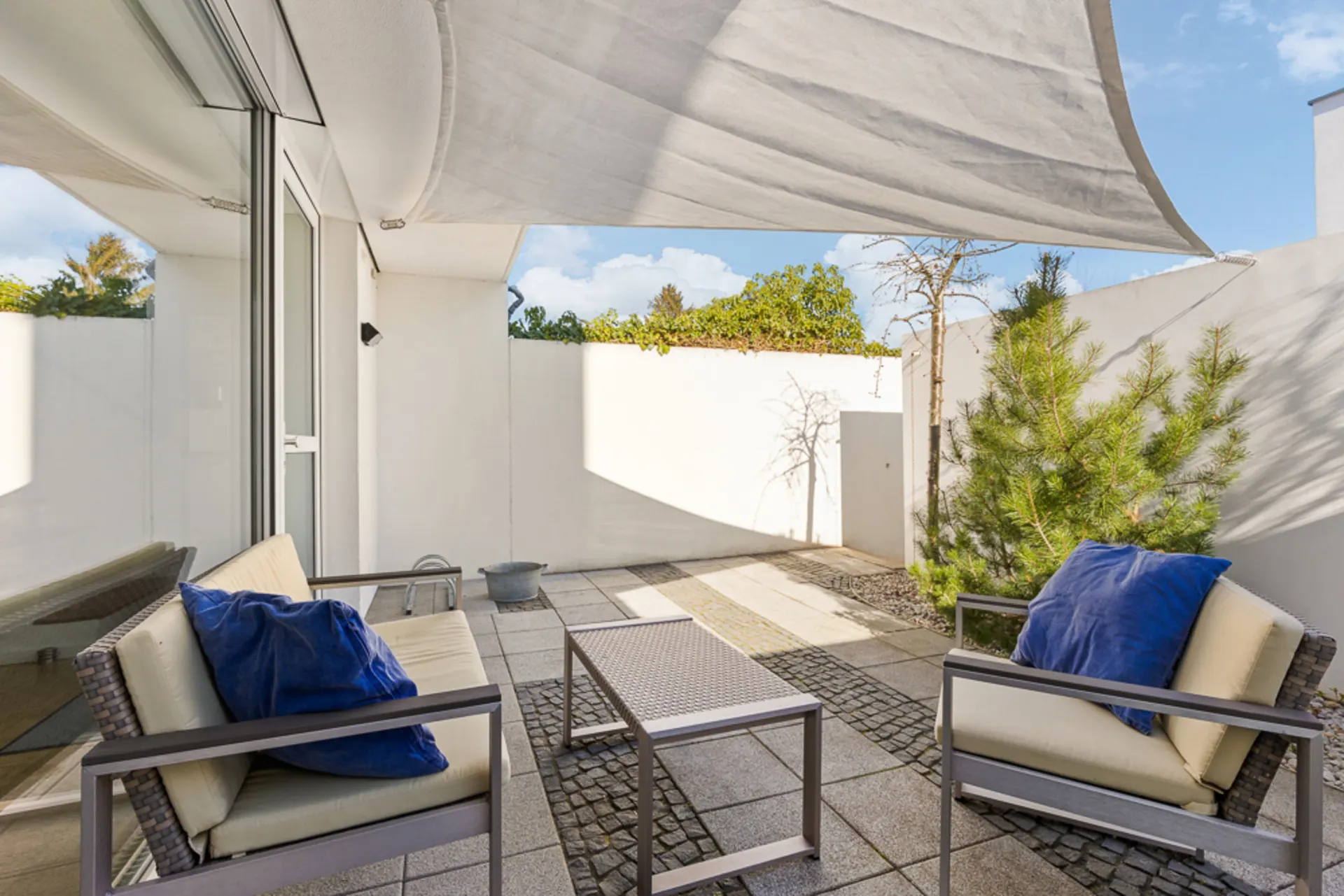Moderne Terrasse mit Sitzmöbeln, Sonnensegel, Kissen und grünen Pflanzen an weißer Mauer.