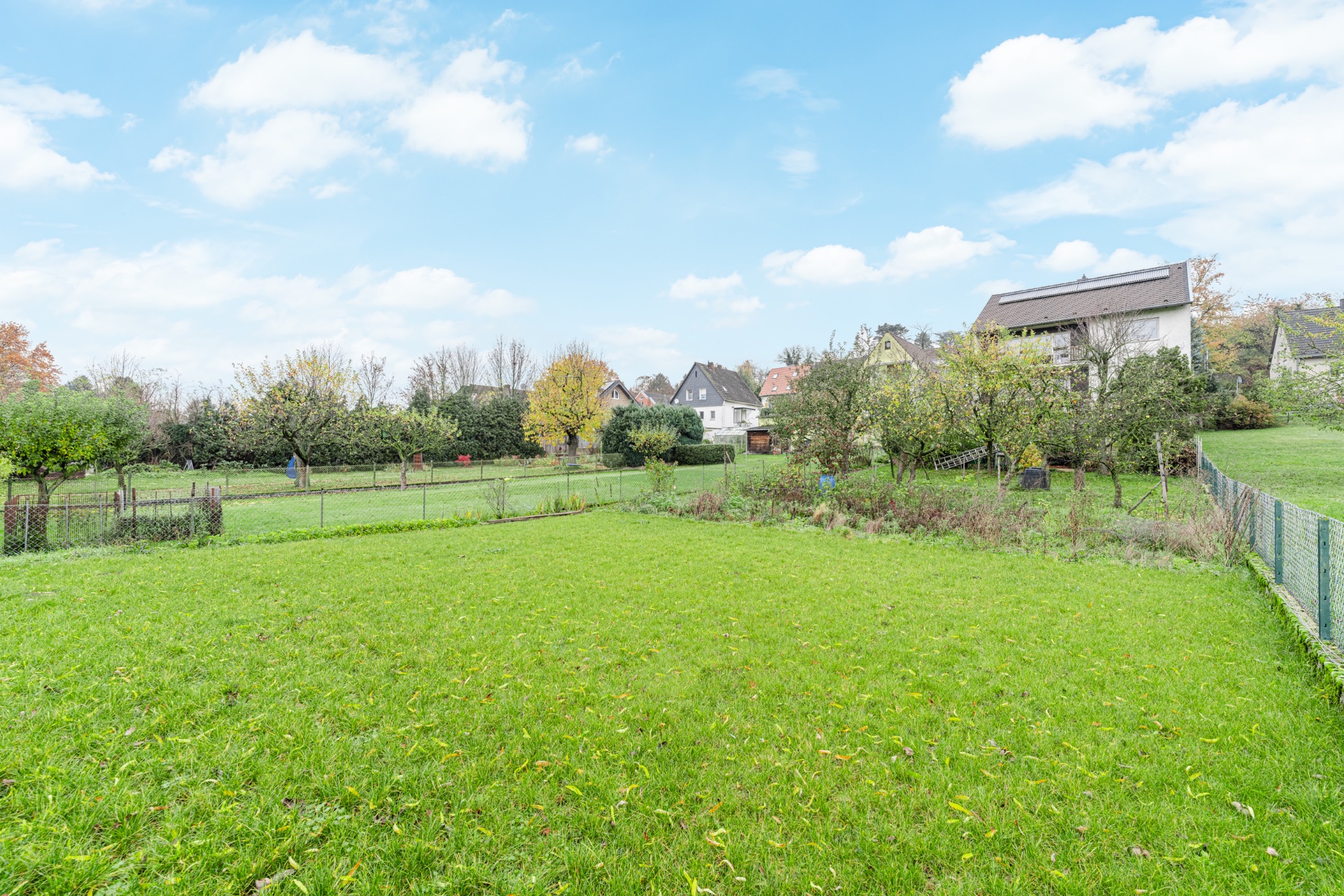 Großer Garten mit Rasenfläche, Bäumen und Blick auf benachbarte Wohnhäuser bei blauem Himmel.