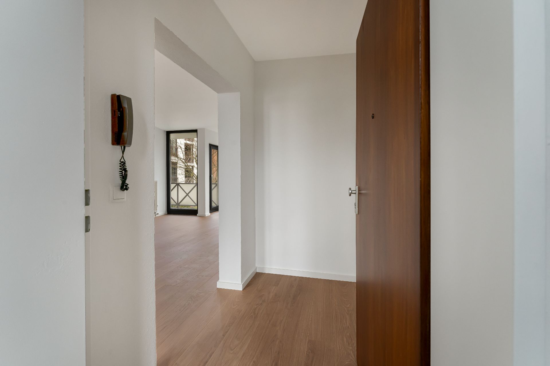 Eingangsbereich einer Wohnung mit Holztür, Flur und Blick in ein helles Zimmer mit großem Fenster.