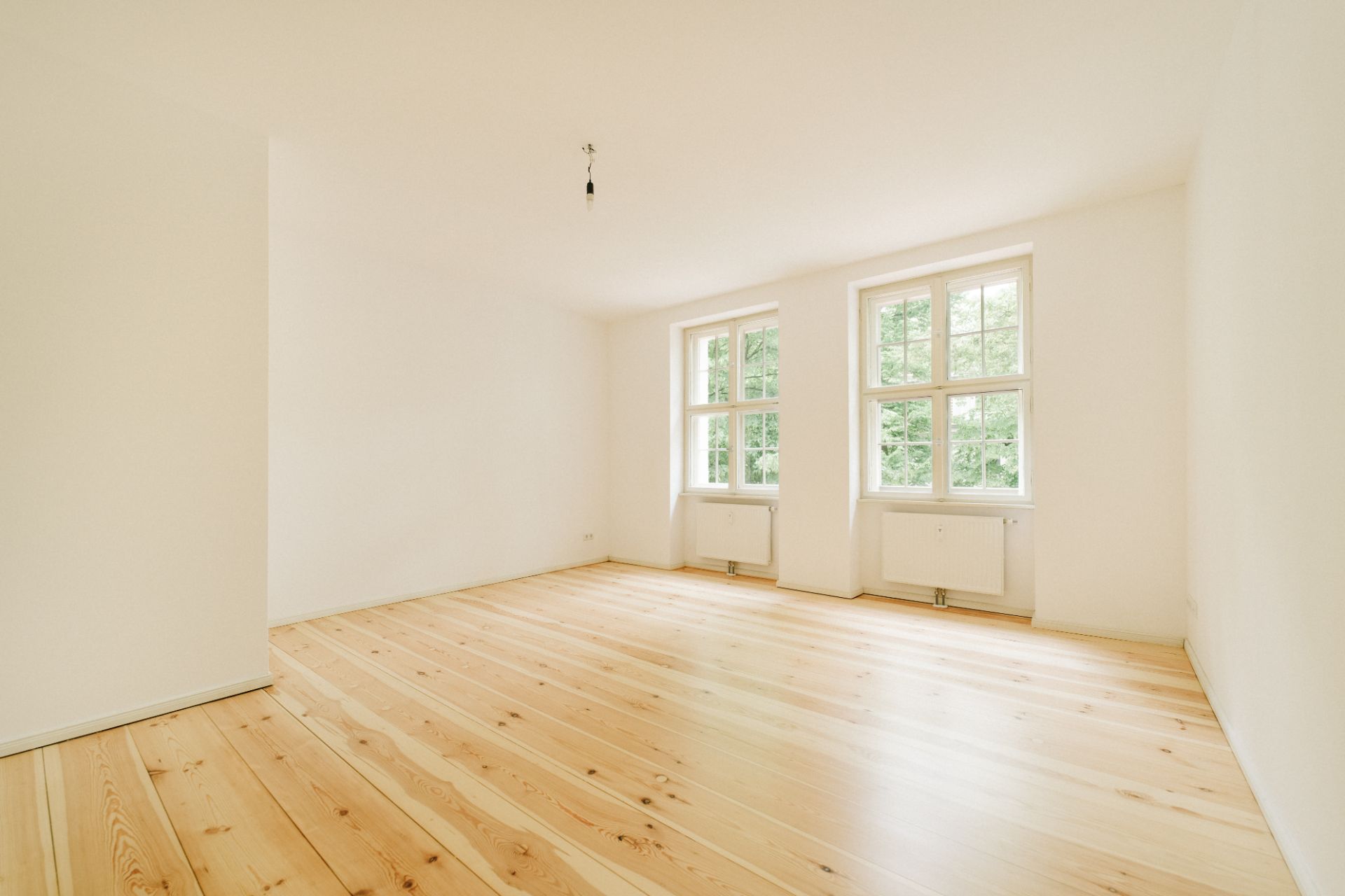 Helles, leeres Zimmer mit Holzboden und zwei großen Fenstern.