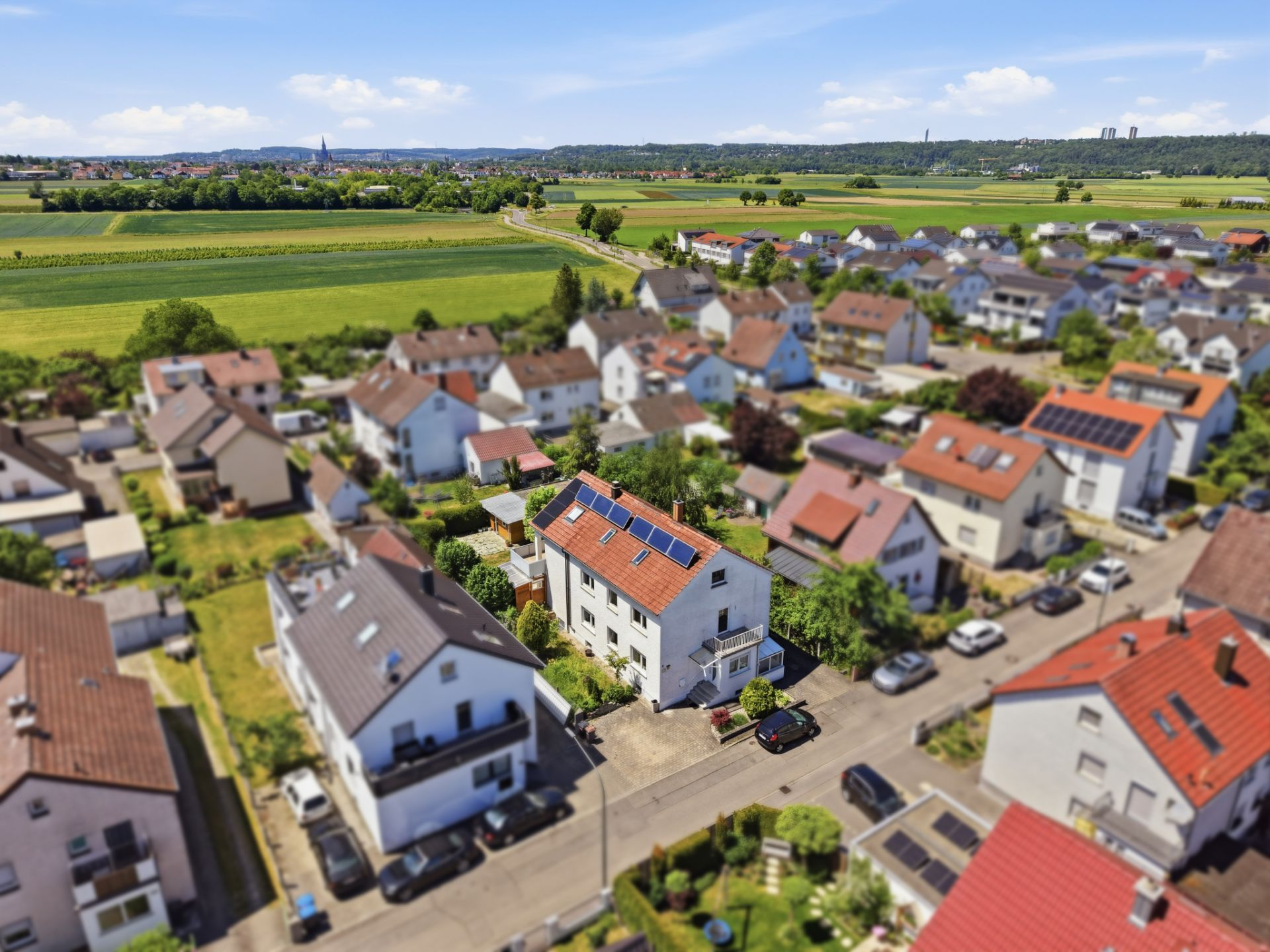 Luftaufnahme eines Wohnviertels mit Einfamilienhäusern und grünen Feldern im Hintergrund.