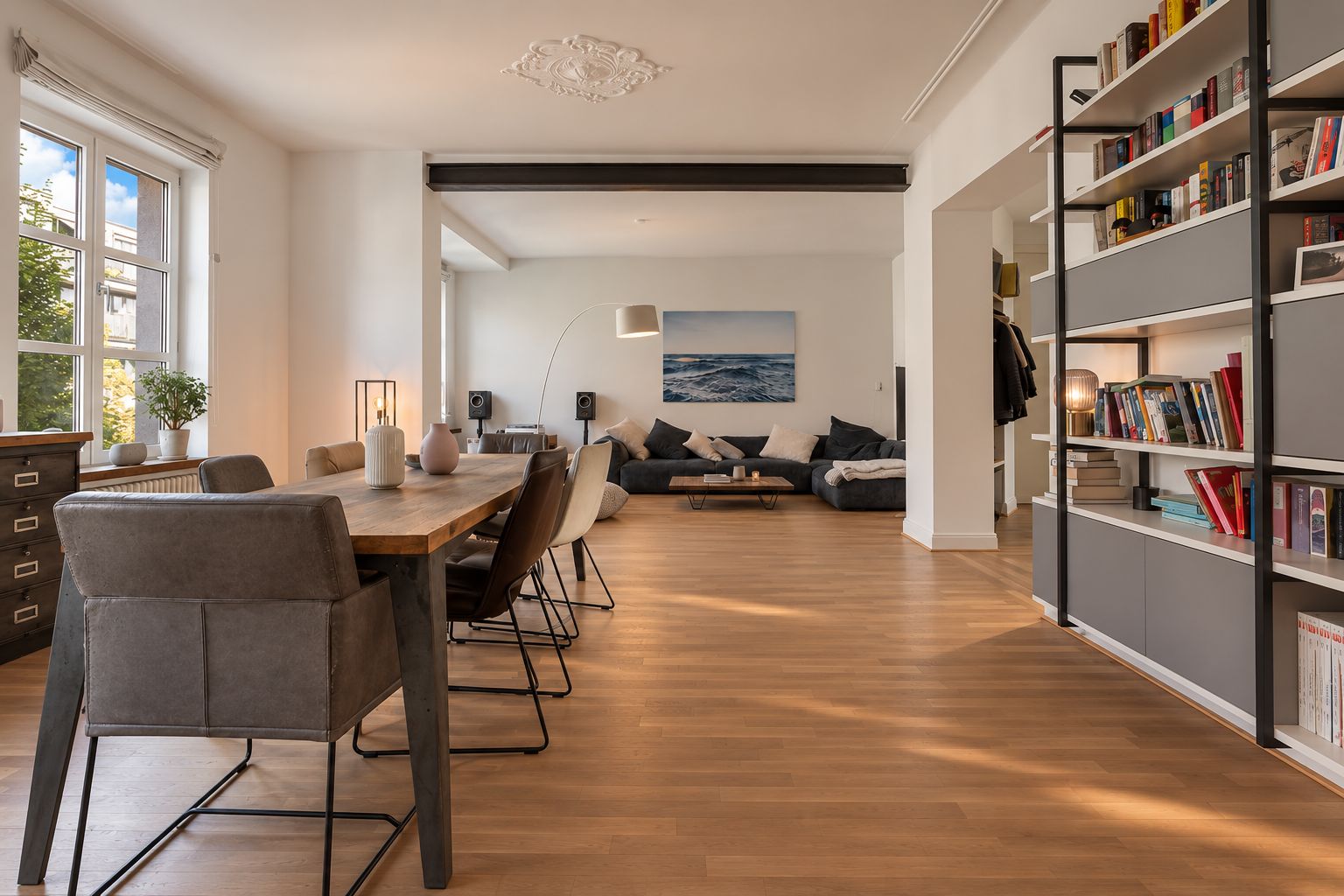 Helles Wohnzimmer mit Esstisch, Bücherregal, Sofa und großen Fenstern.